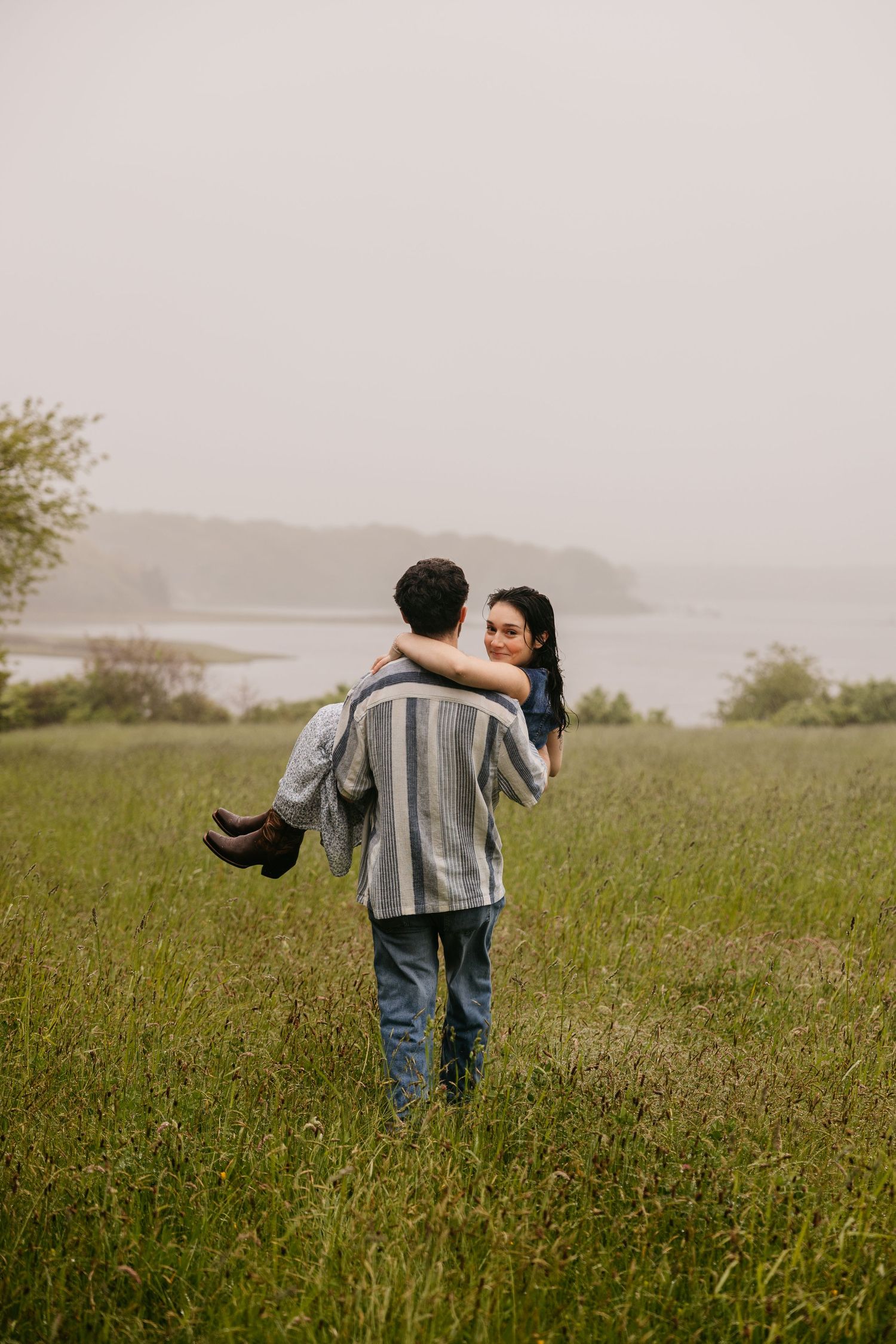 A couple shares a romantic moment in a misty green field, the man carrying the woman in his arms near a coastal landscape.