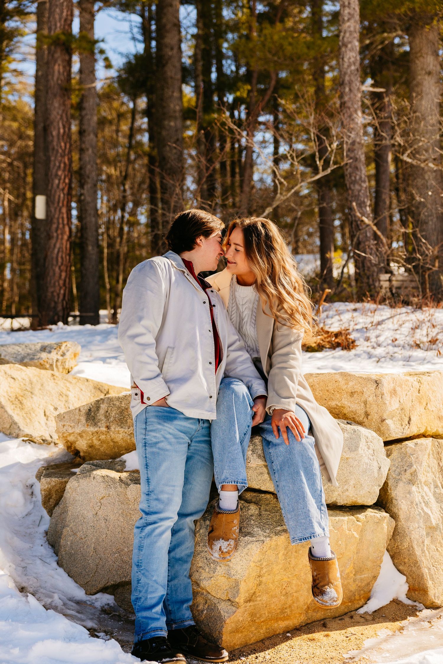 Couple sharing a kiss on snowy rock wall surrounded by pine trees in winter, wearing jeans and casual jackets.