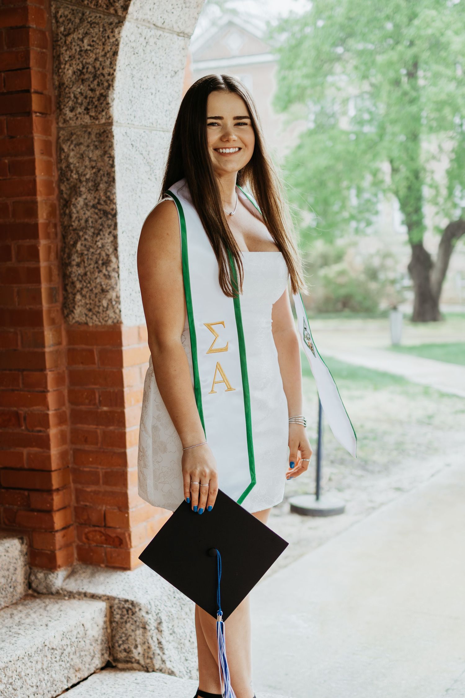 Graduate in white dress and green stole posing with diploma near brick columns.