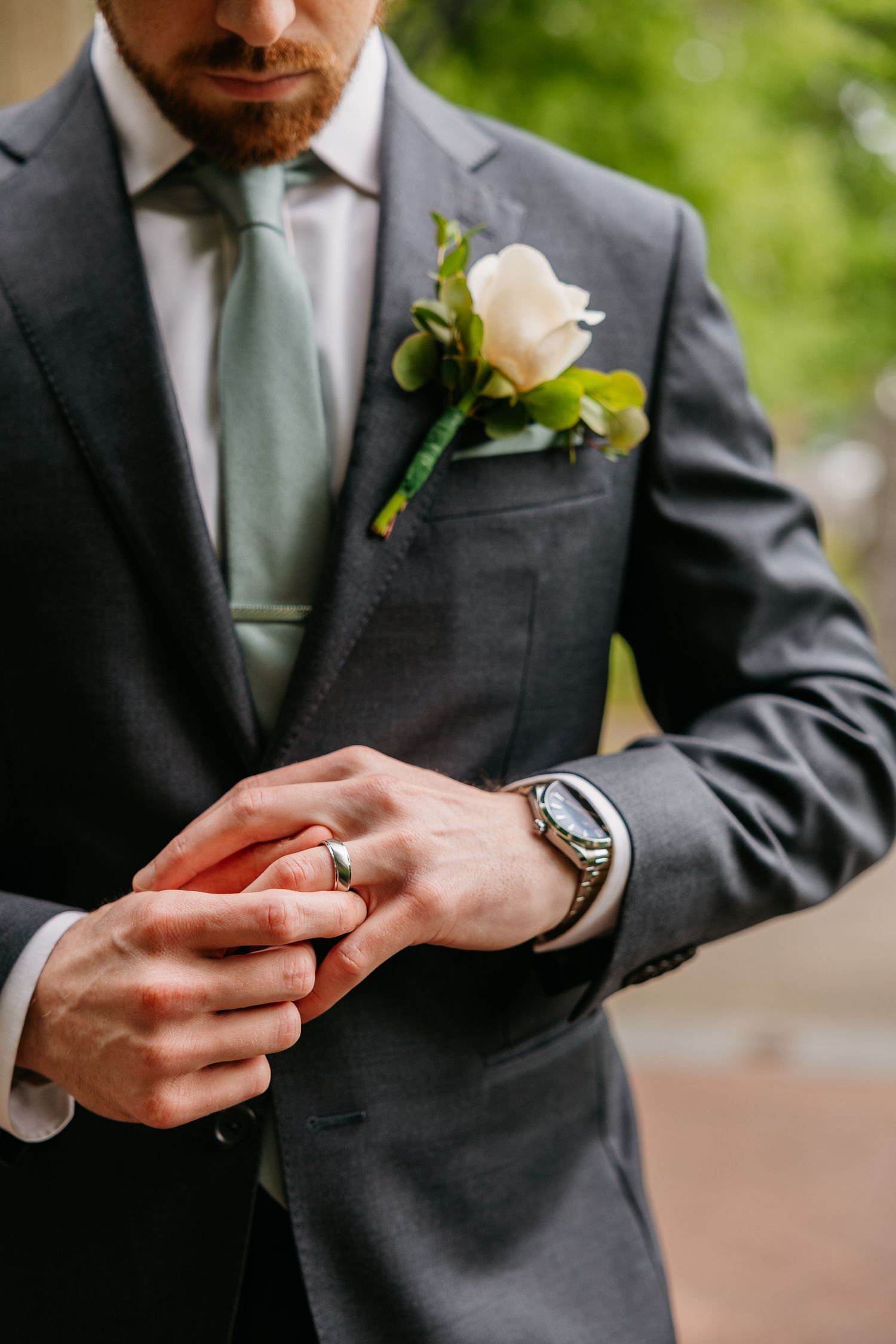 Groom in gray suit with green tie and white boutonniere adjusts wedding ring on finger, wearing elegant watch.