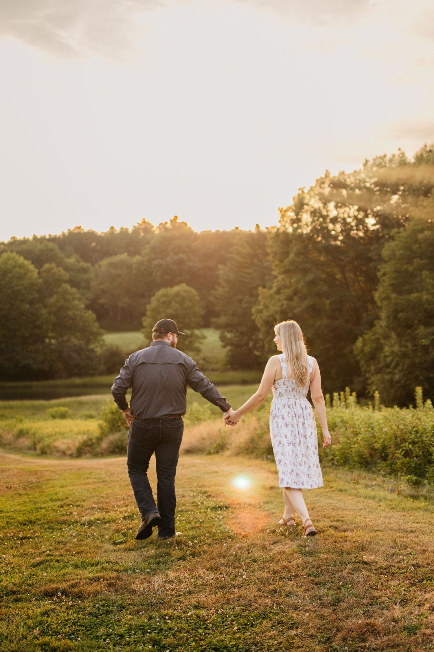 A couple walks together in a grassy meadow at sunset with golden light filtering through trees in the background.