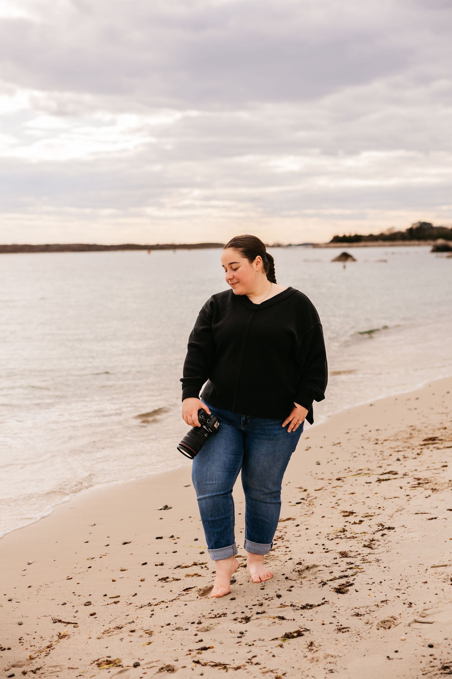 Woman in black sweater and jeans walks barefoot on sandy beach at sunset, holding a camera.