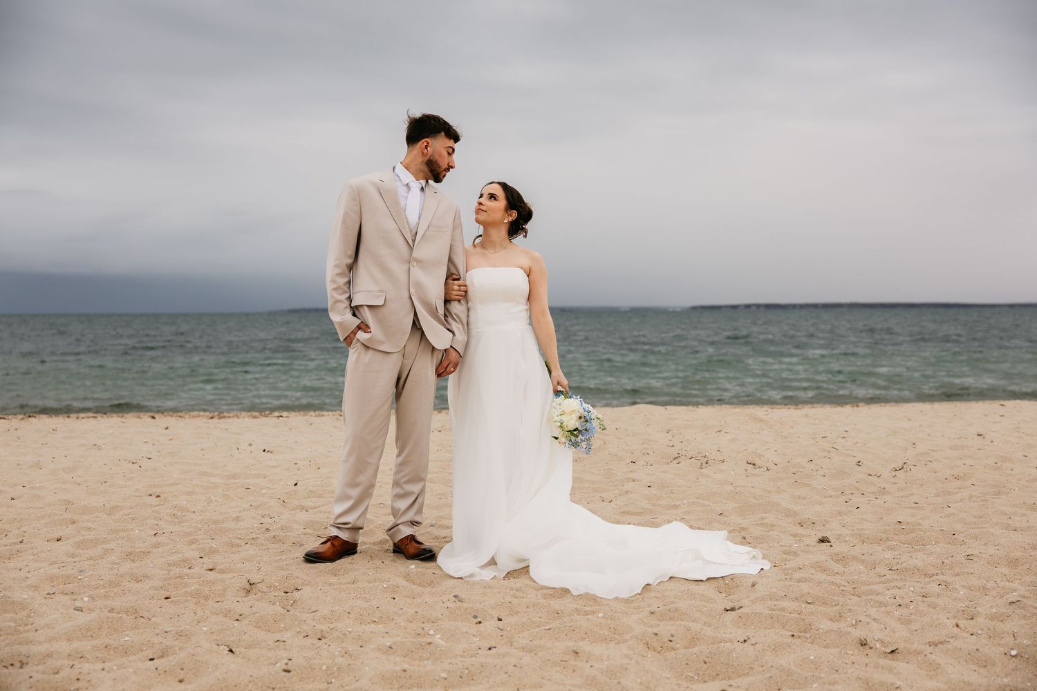 Bride and groom holding hands on sandy beach, wearing white gown and tan suit, with ocean backdrop on cloudy day.