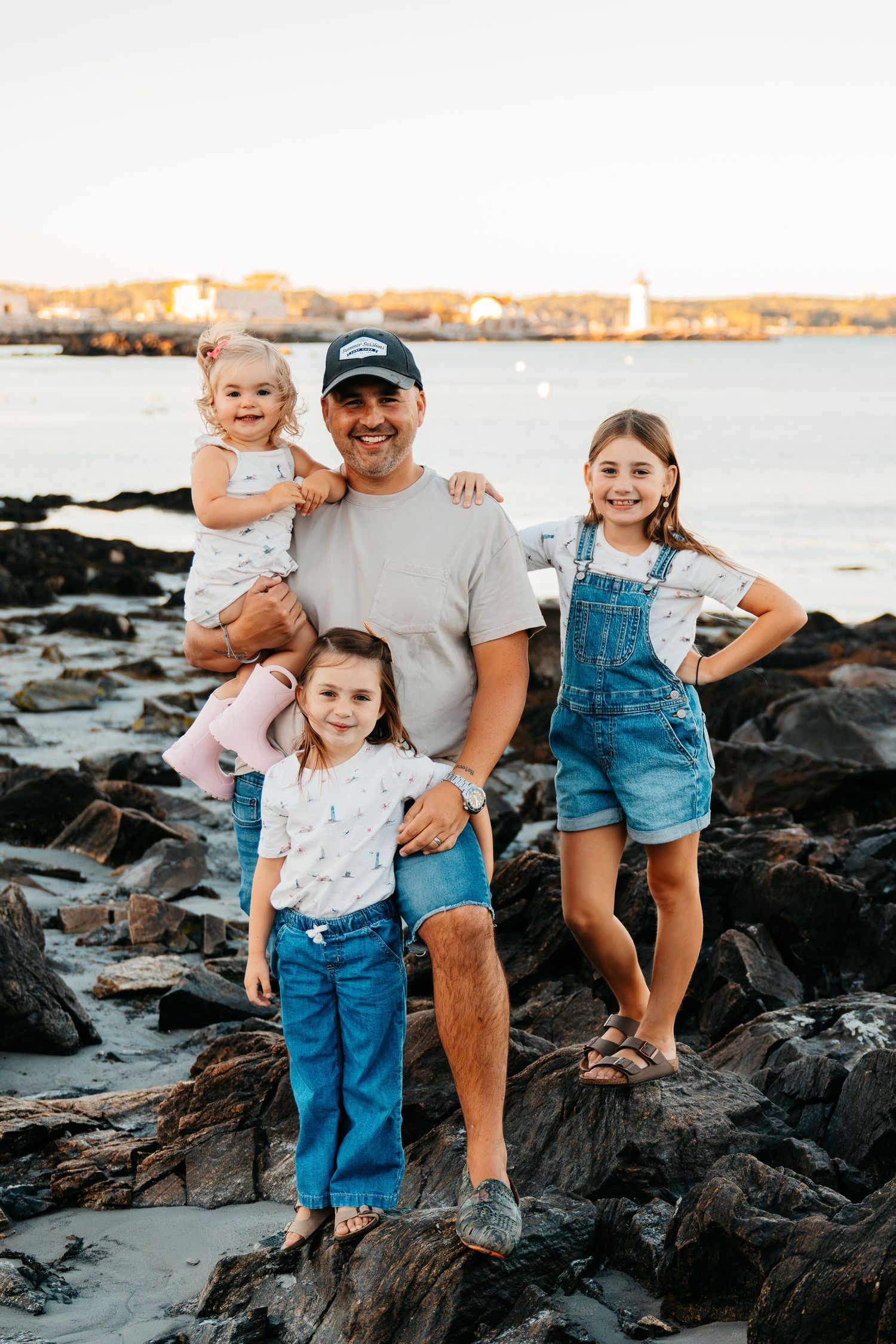 A family poses together on rocky coastal shoreline at sunset with water and distant shore in background.