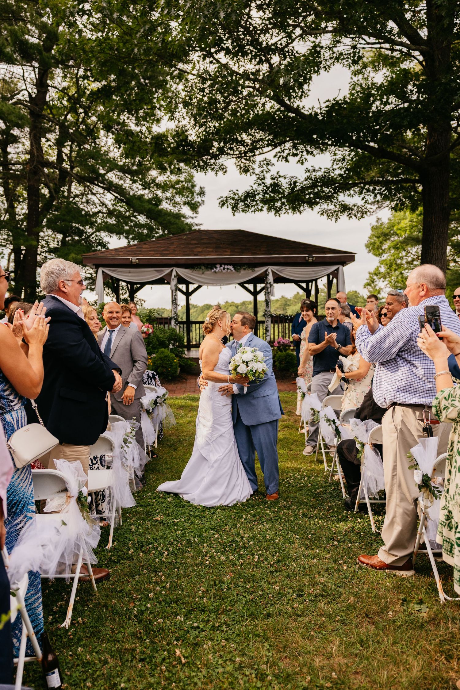 Bride and groom share a kiss at outdoor wedding ceremony aisle, guests applauding, gazebo backdrop.
