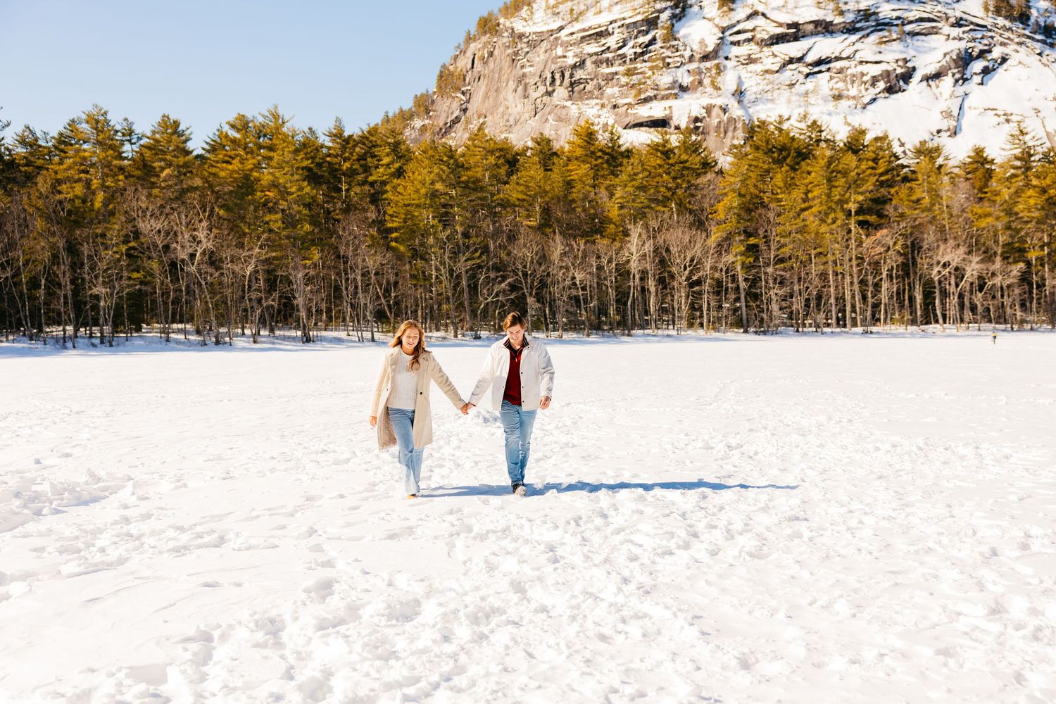 Couple holding hands walking on snowy field with pine trees and rocky cliff in sunny winter landscape.
