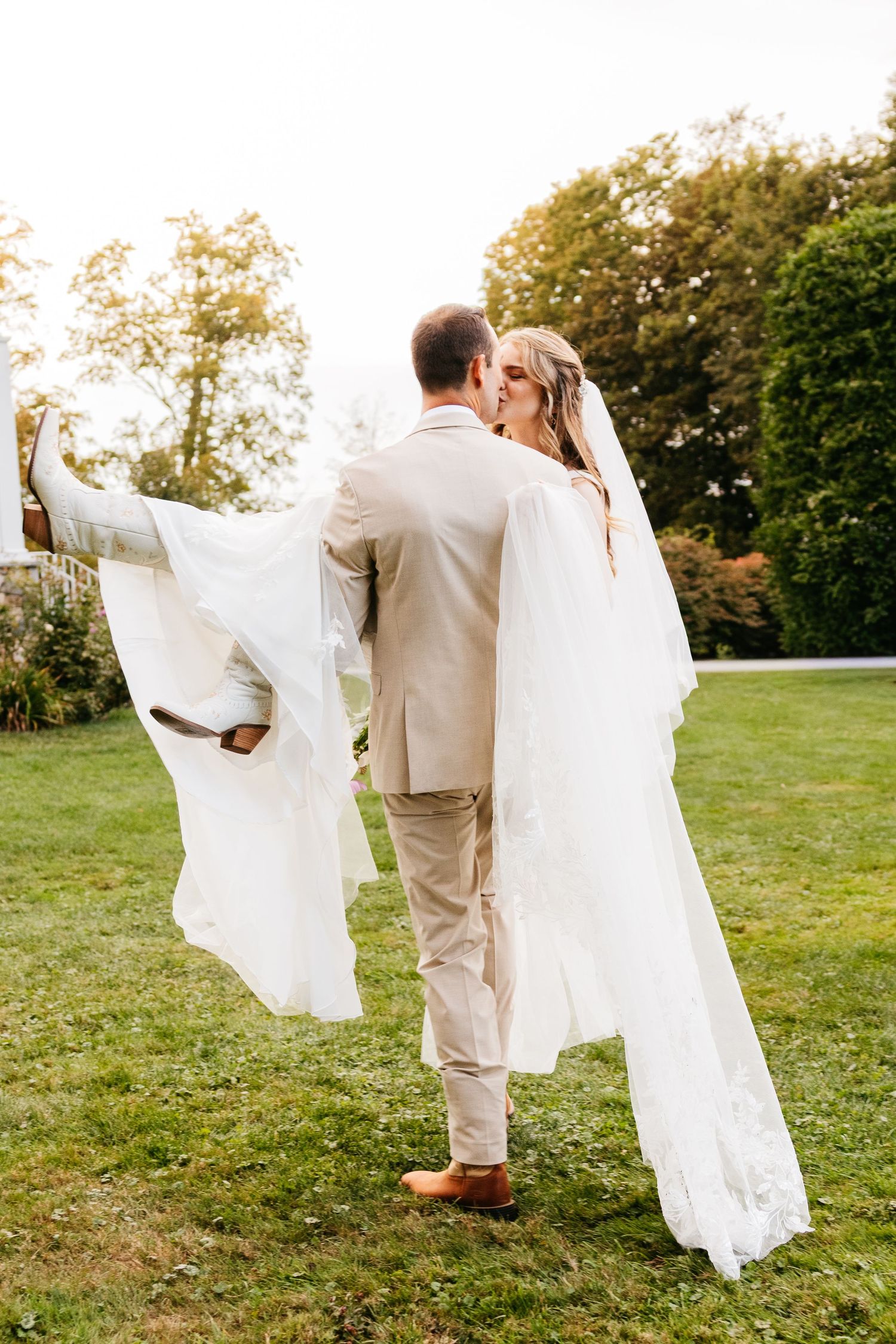 A couple in wedding attire dances together on a grassy hill during golden hour, with the bride's dress flowing in the breeze.
