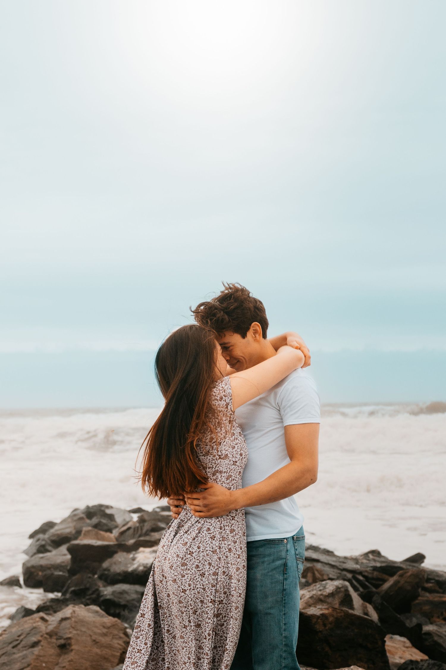 A romantic couple embraces on a rocky beach against a cloudy sky backdrop.