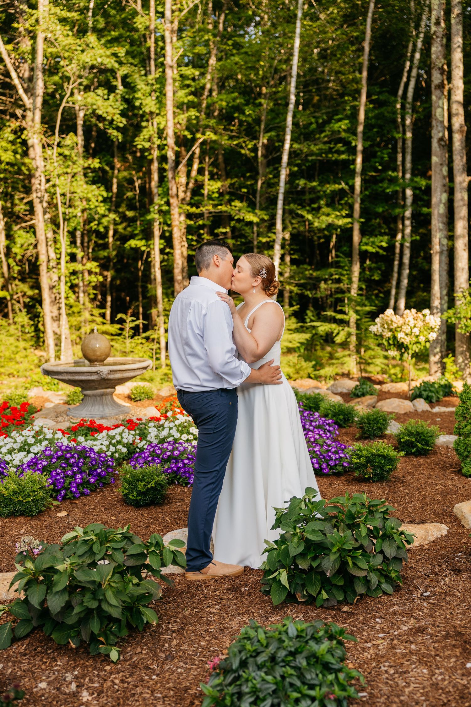 A couple shares a romantic kiss in a colorful garden with red, purple, and white flowers, surrounded by lush green trees.