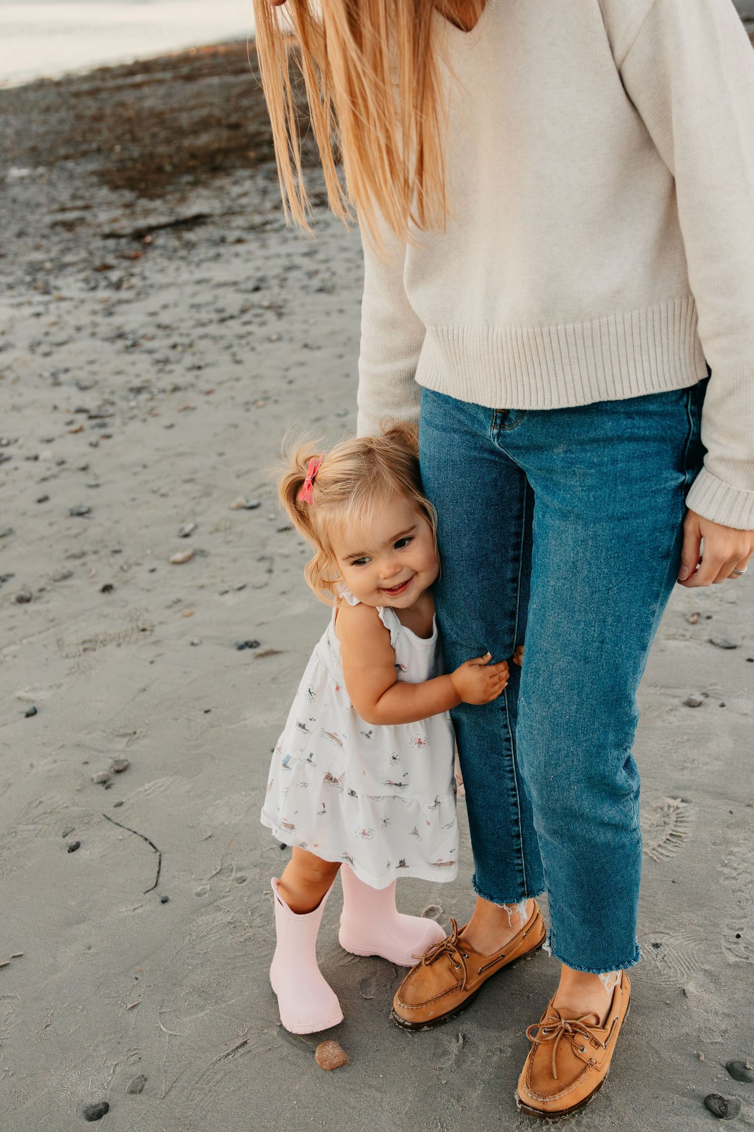 A small child in a white dress hugs an adult's leg while standing on a beach wearing pink socks.