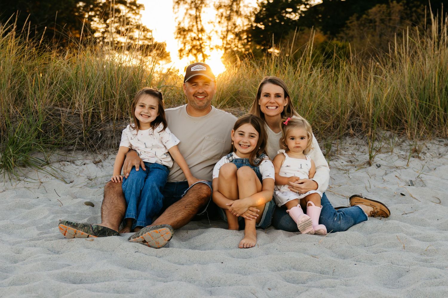 Family of five smiling together on sandy beach at golden sunset with tall grass in background.