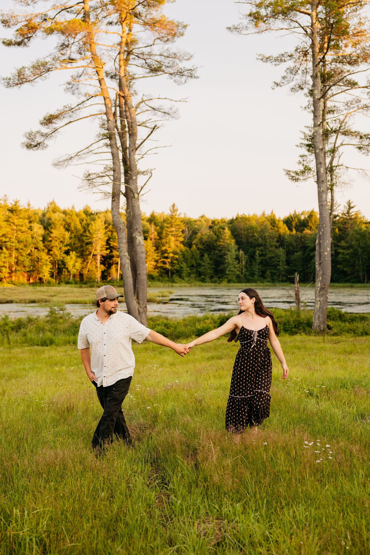 Couple holding hands walking through a sunlit green meadow with tall pine trees and a golden forest at sunset.