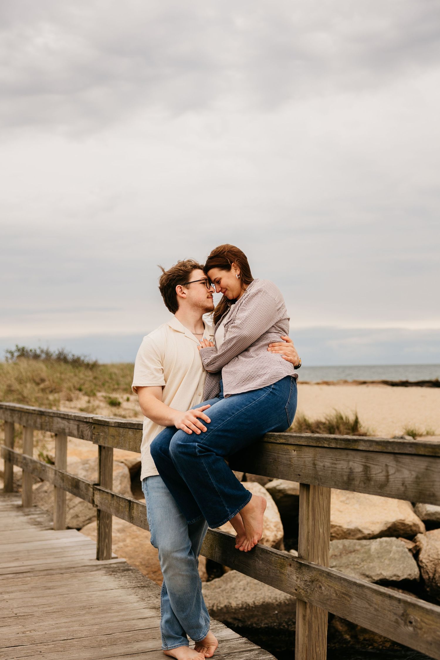 Couple sharing a romantic moment on a beach boardwalk, woman sitting on railing as man holds her close.