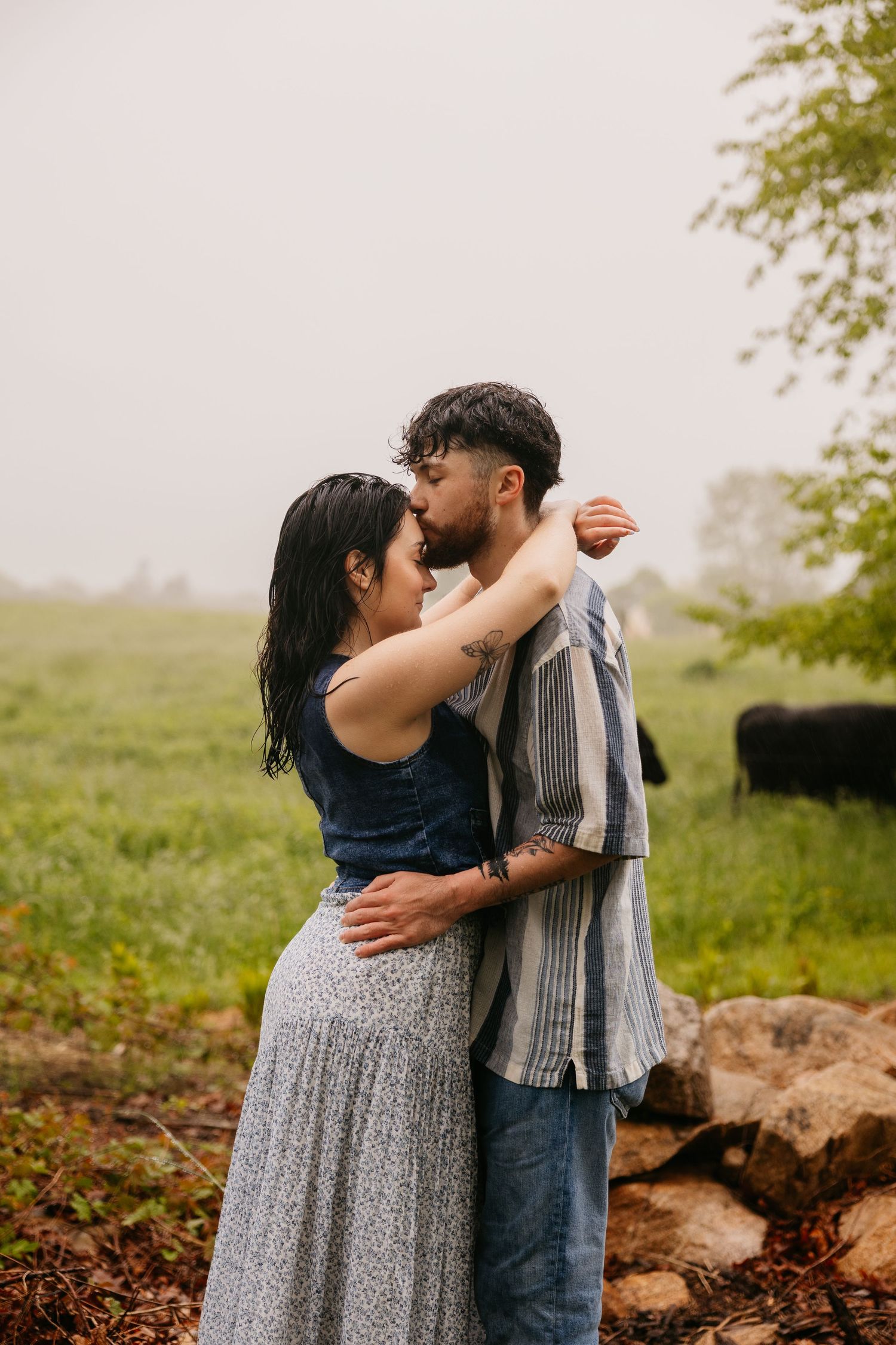 A couple embraces romantically in a misty green field, the man kissing her forehead while she wraps her arms around him.