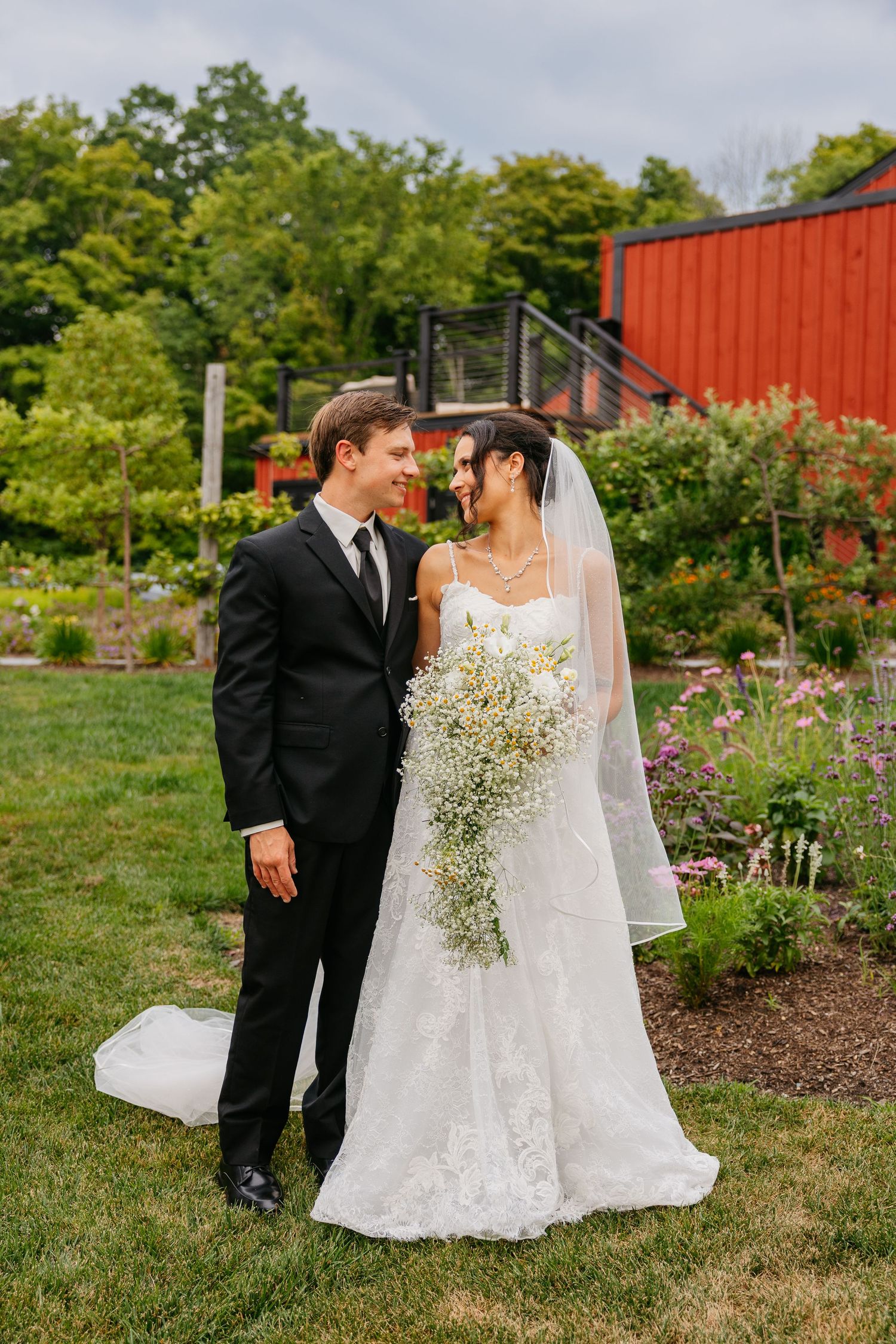 Bride and groom share a romantic moment in a garden, she holds a baby's breath bouquet, wearing lace gown and veil.