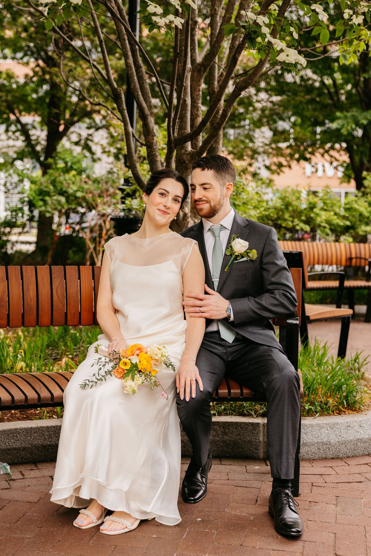 A bride and groom sit together on a park bench, she holding a yellow bouquet in a white gown, he in a gray suit.