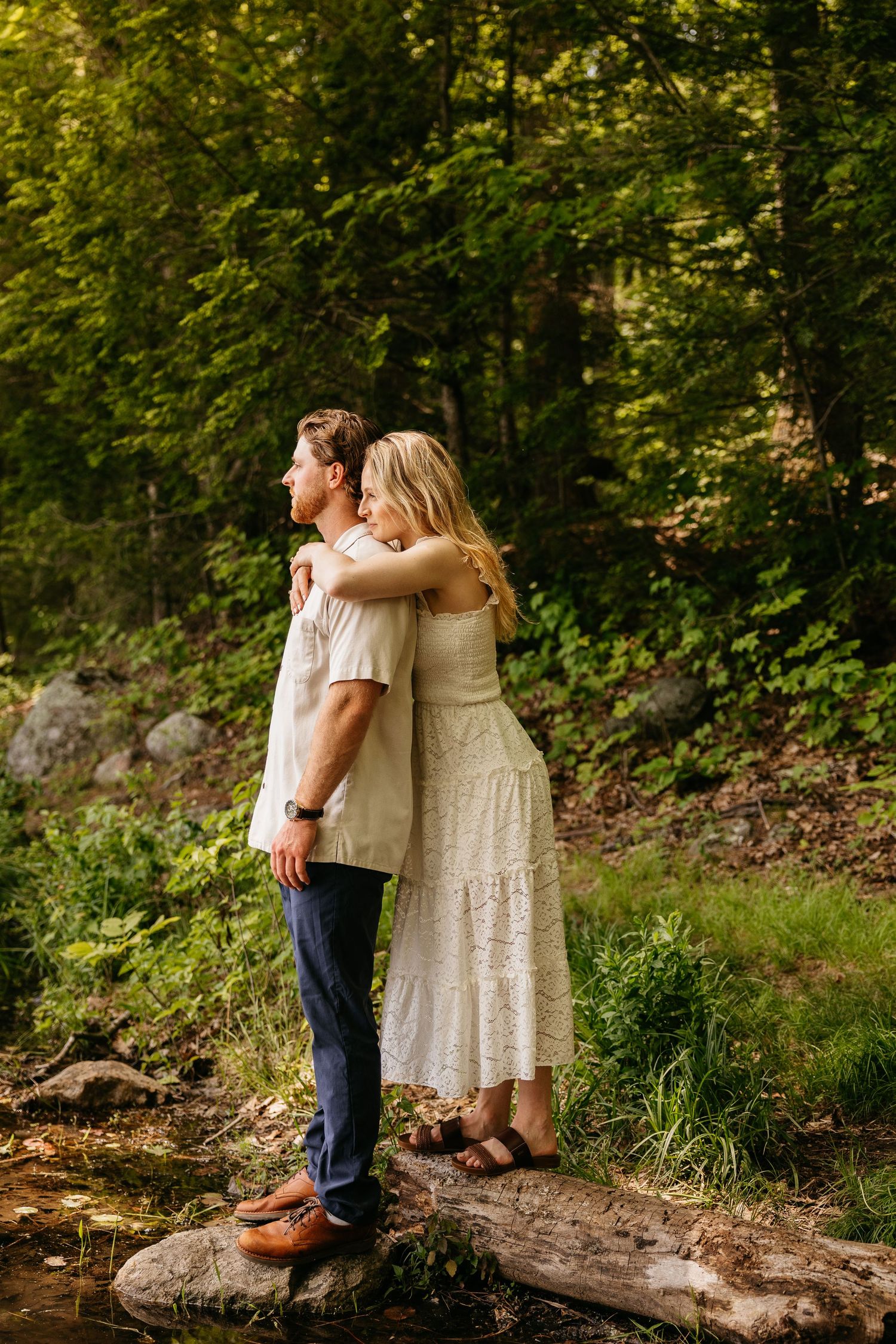 Couple embracing in a lush forest, woman in white lace dress hugging man from behind while standing on rocks near a stream.