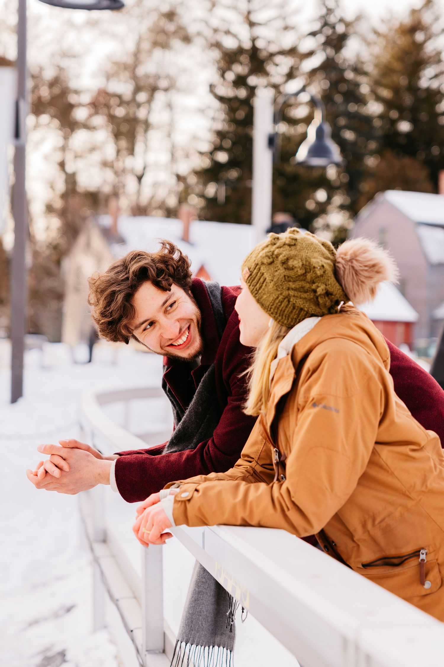 Two people share a playful moment leaning against a snow-covered railing in winter.
