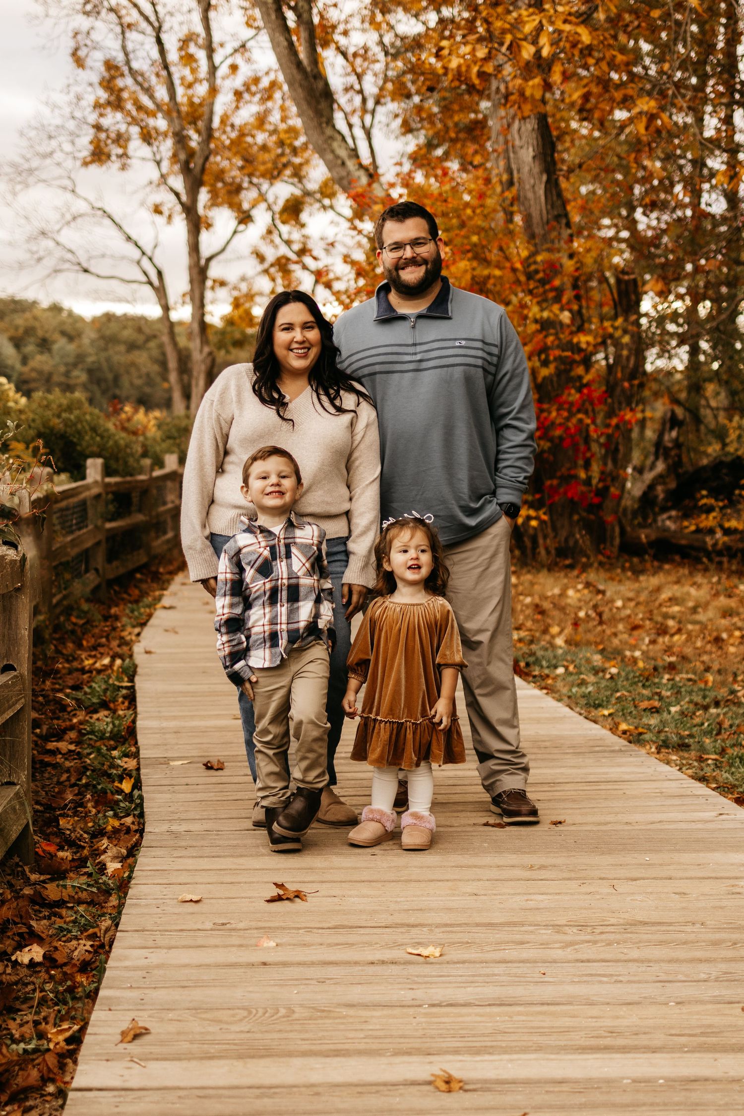 Family of four posing on a wooden boardwalk surrounded by colorful autumn foliage for a fall portrait.