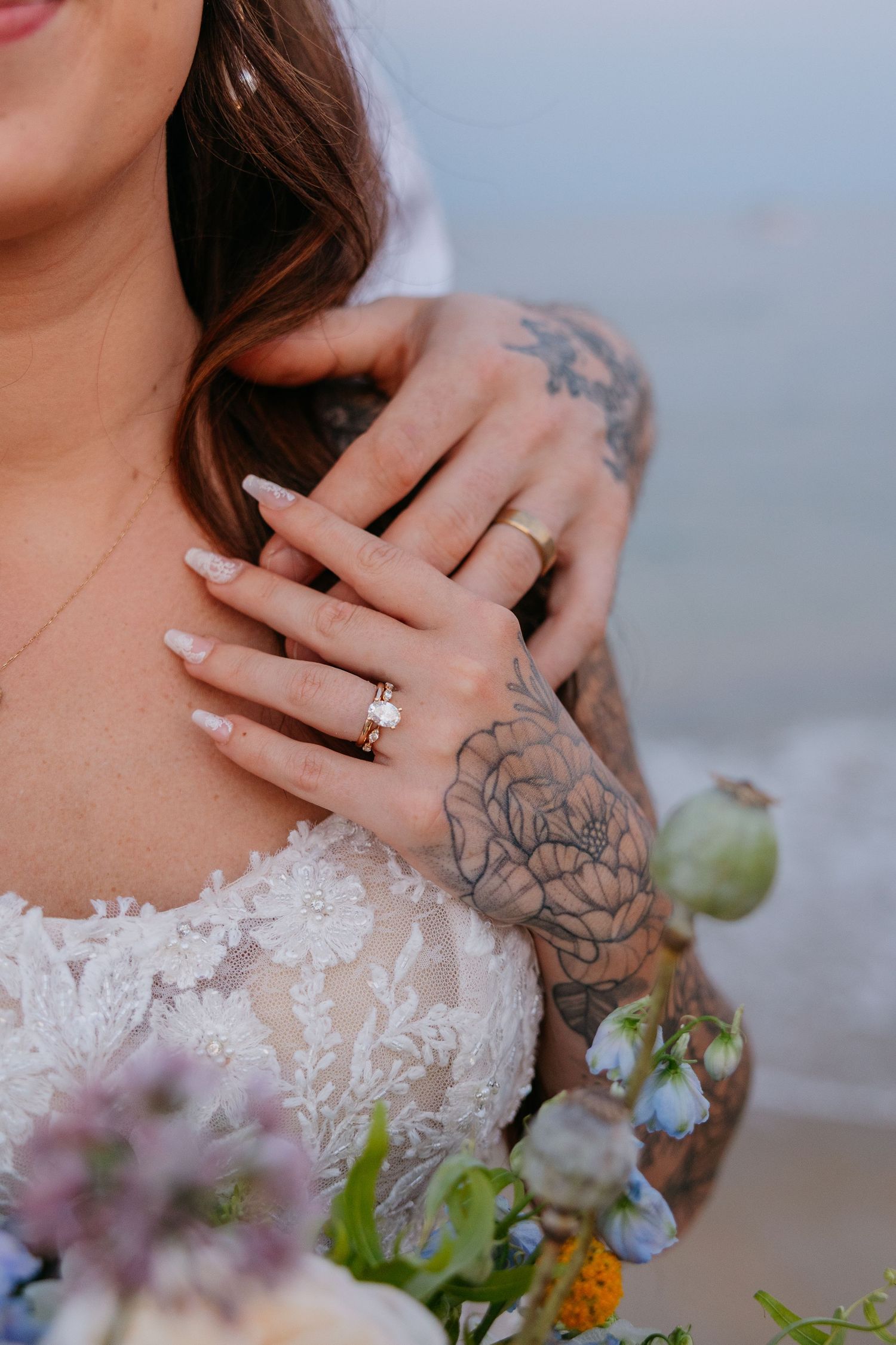 Close-up of a bride in lace wedding dress with tattooed arms, displaying engagement ring and wedding band while holding a floral bouquet.