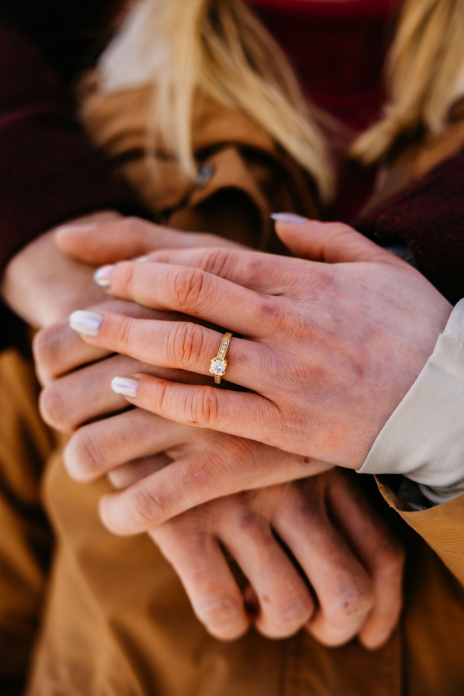 Close-up of intertwined hands showing a gold diamond engagement ring on a woman's finger with white nail polish.