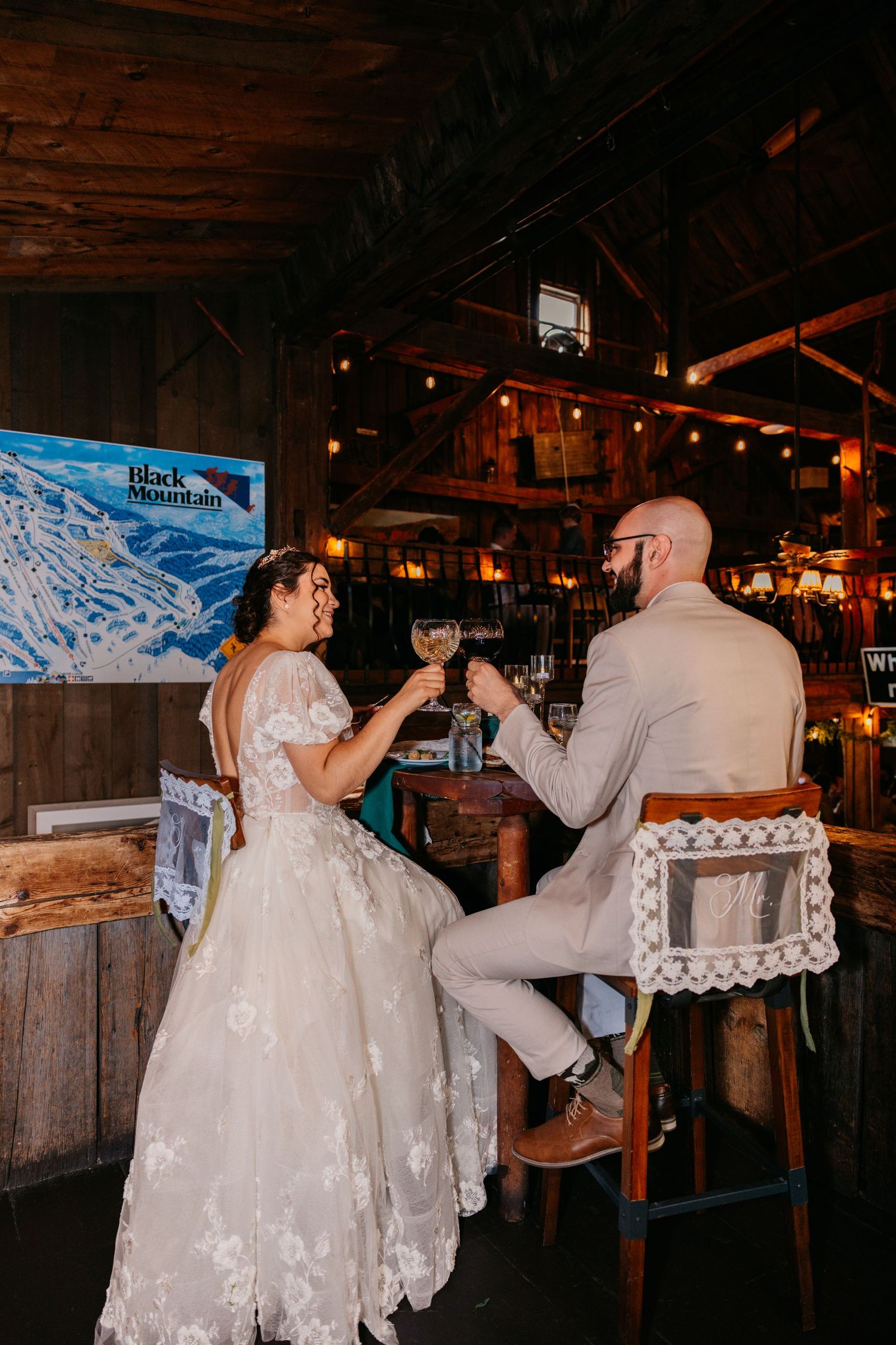 Bride and groom toast with wine glasses at a rustic bar, wearing wedding attire inside a warmly lit wooden venue.