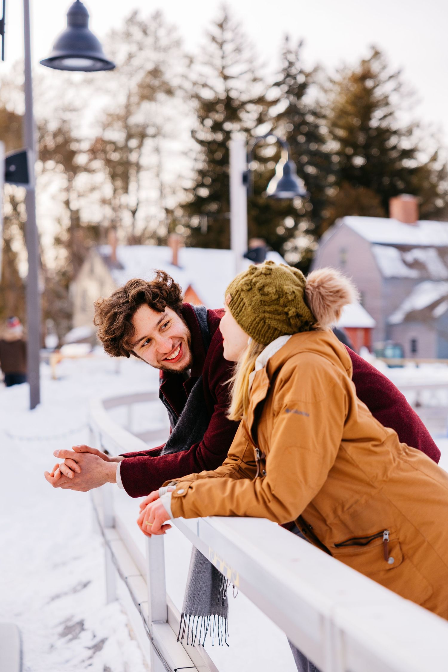 A smiling couple leans on a white railing in a snowy winter setting, with the woman wearing a yellow coat and knit hat.