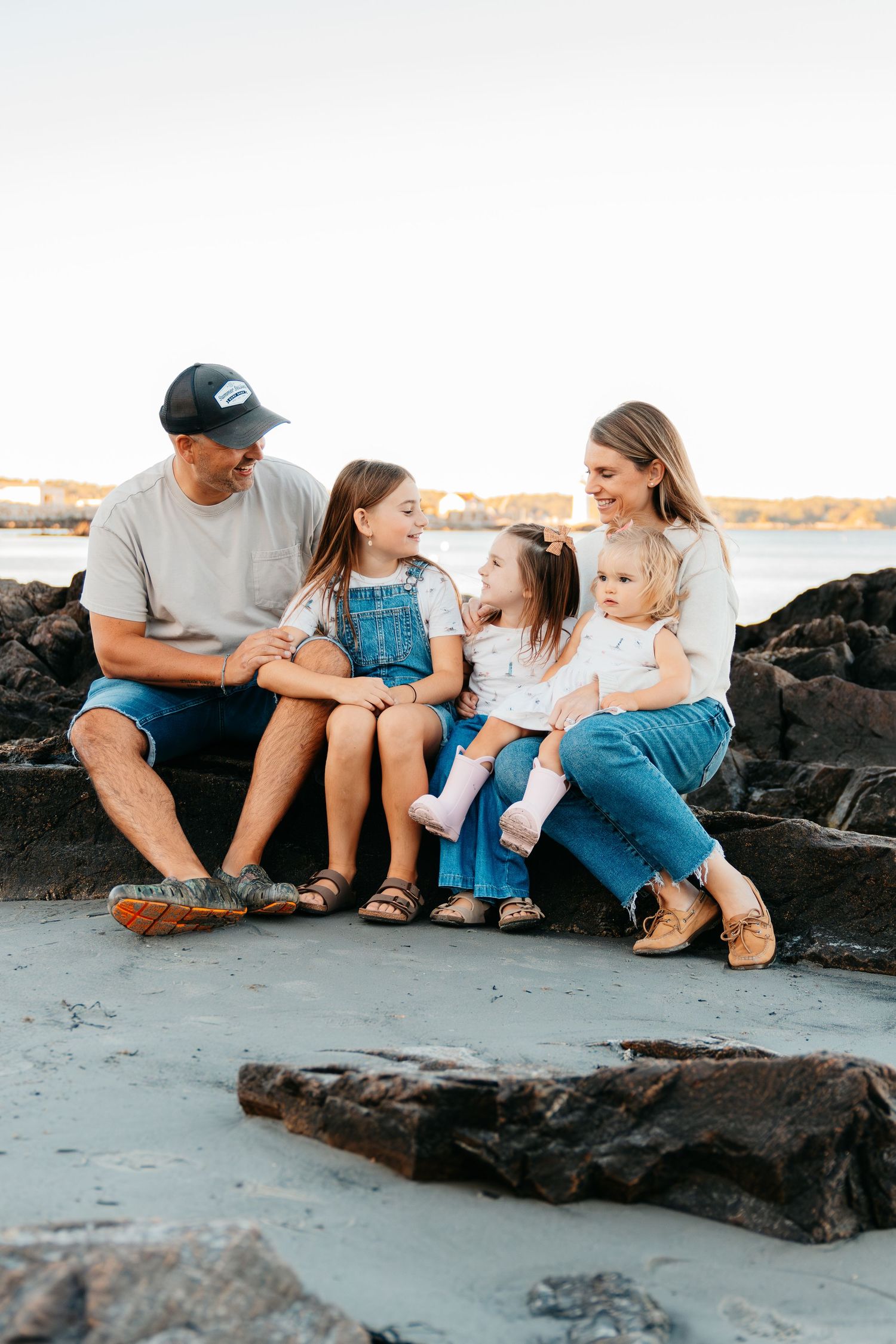 Family of five sitting on rocks at the beach, two parents smiling with three young daughters in denim outfits.