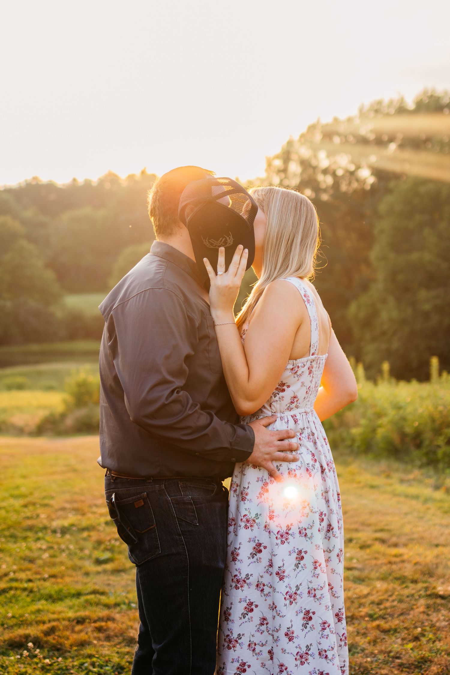 Couple sharing a romantic kiss at golden hour sunset in a lush green countryside field, woman in floral dress.