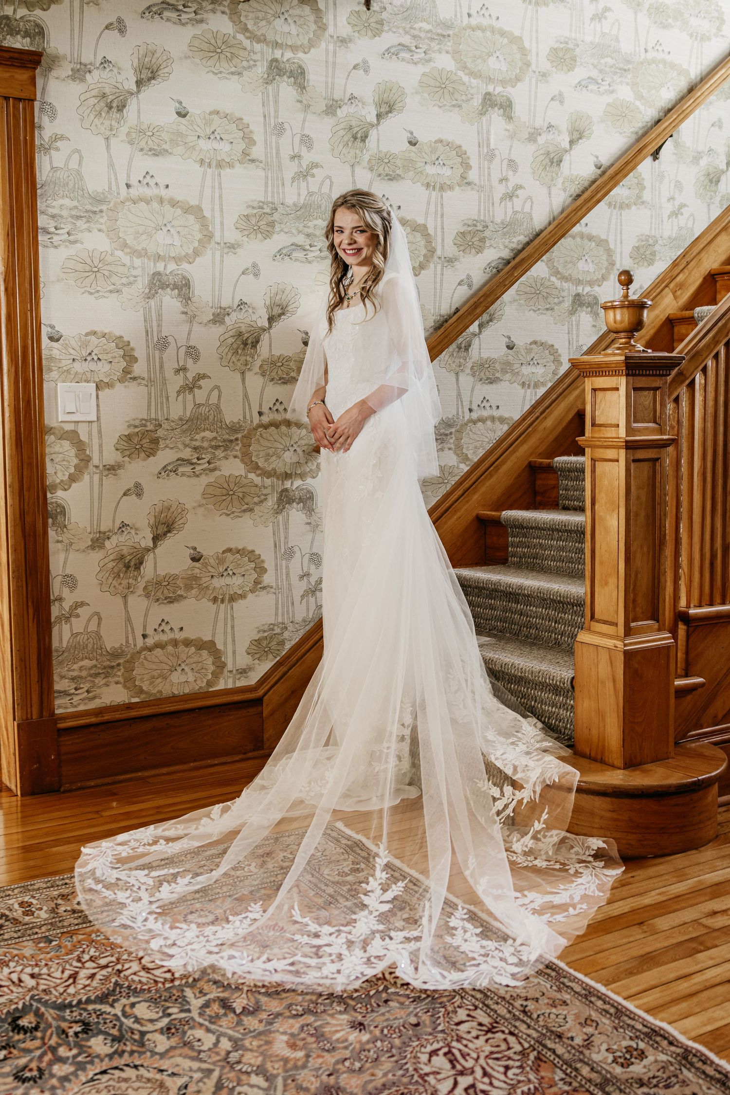 Wedding dress train and veil flowing across hardwood floor with vintage wallpaper backdrop.
