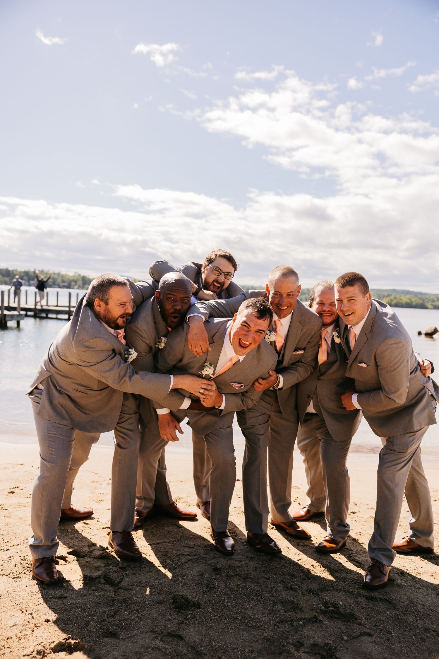 Groomsmen in gray suits laughing and hugging the groom on a sandy beach by a lake on a sunny wedding day.
