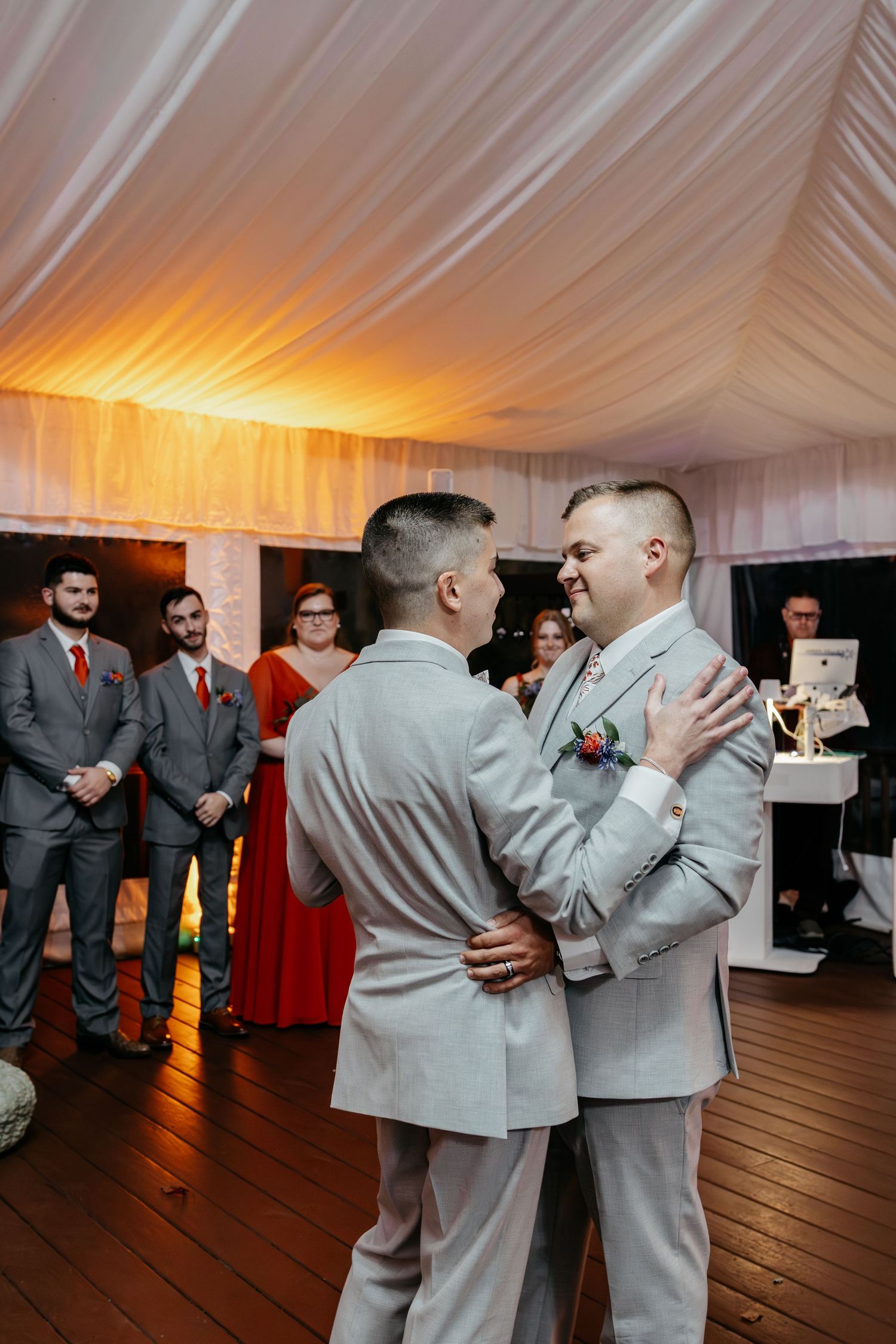 A couple sharing their first dance in light colored suits surrounded by wedding party members.