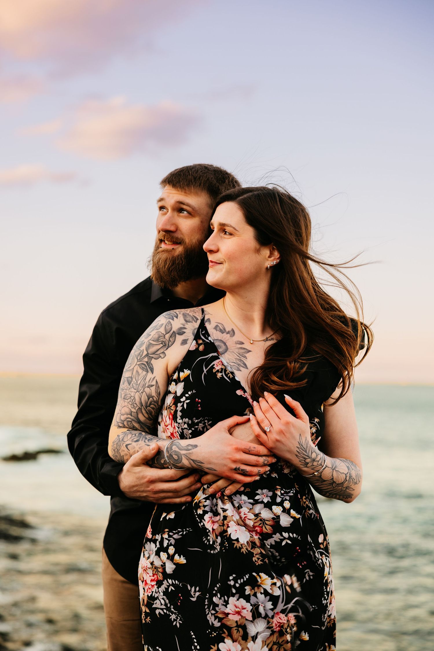 Tattooed couple embracing at sunset beach, woman in floral dress with wind-blown hair, man in black shirt holding her close.