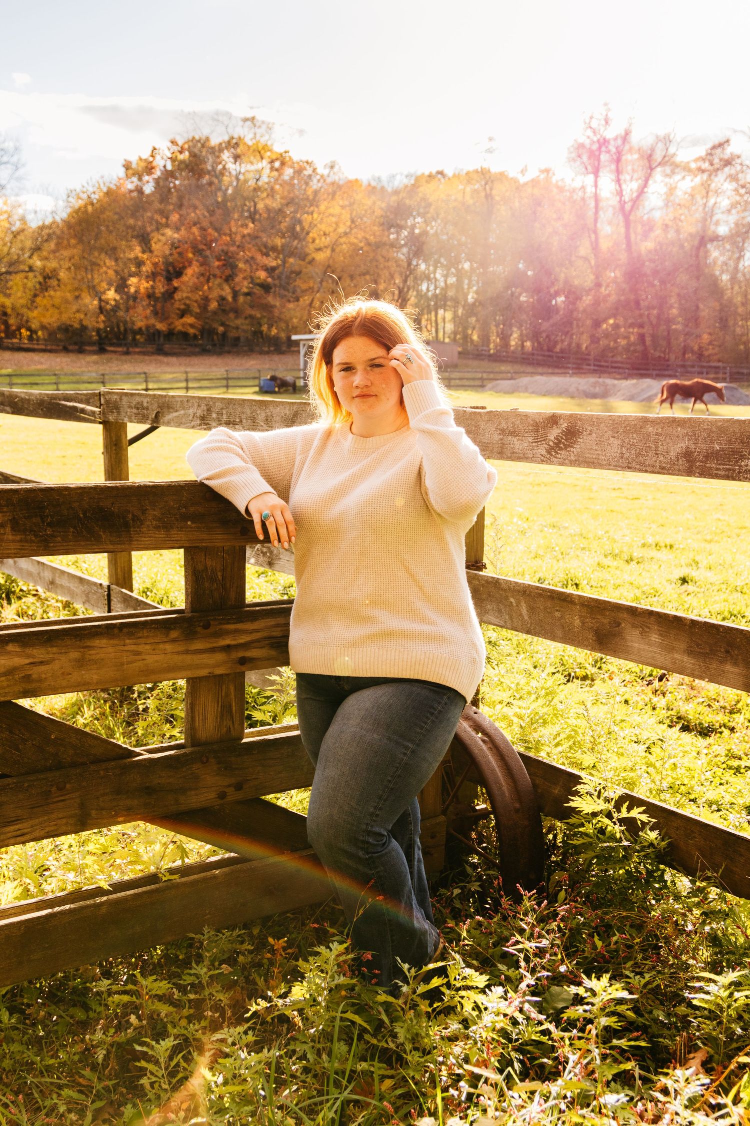 A person in a cream sweater and jeans leans against a wooden fence in a sunlit autumn field.