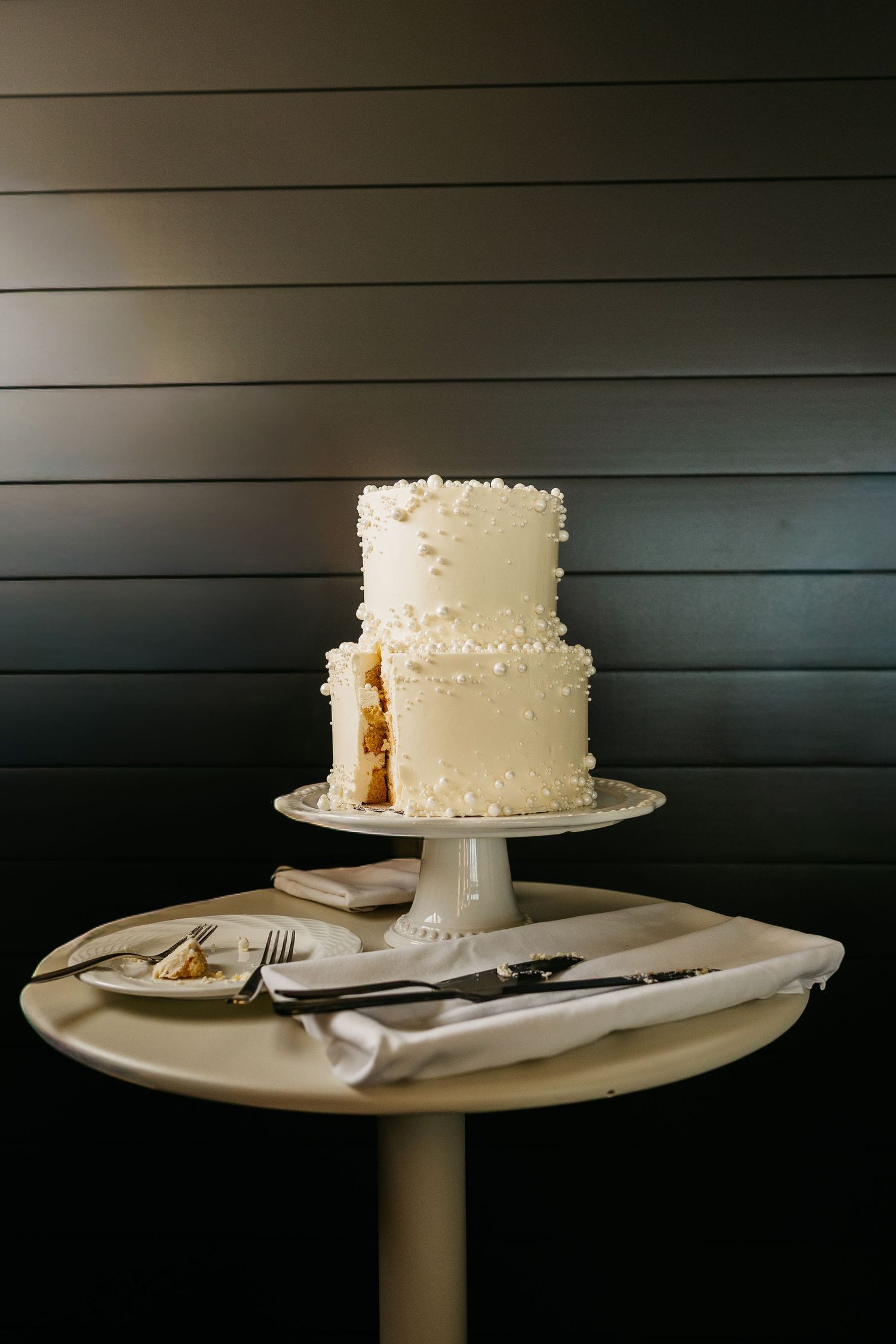 Elegant two-tier white wedding cake with pearl details on a cake stand, displayed on a round table against dark shiplap wall.