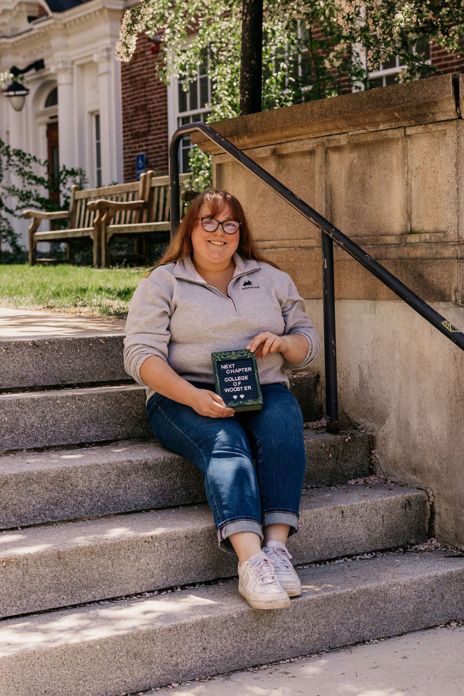 Graduate sitting on concrete steps holding graduation cap in casual pose.