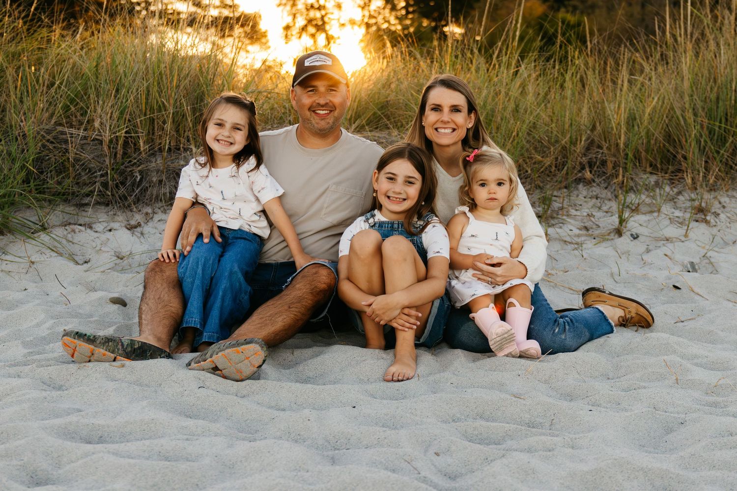 A family sits together on a sandy beach at sunset with grass in the background.