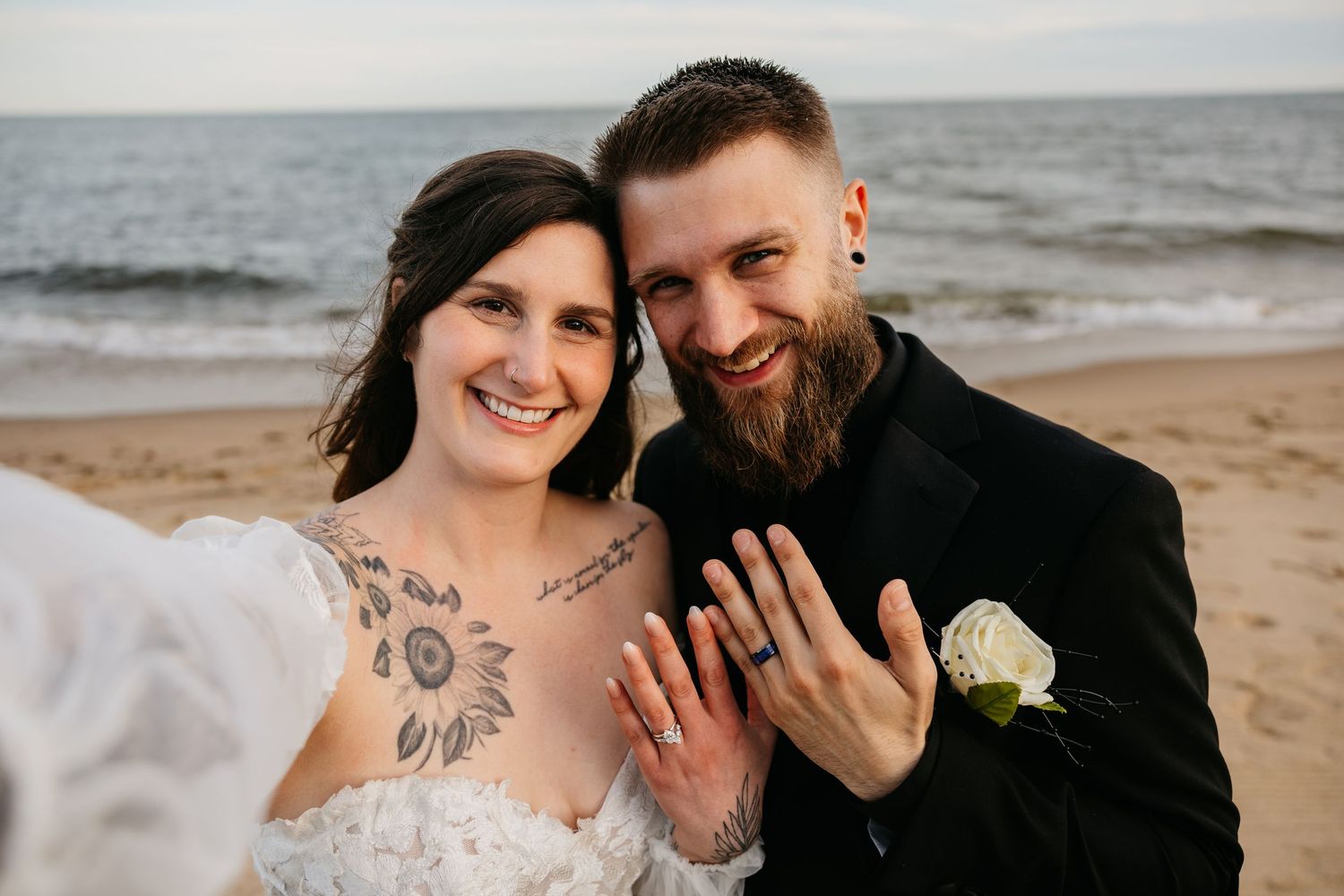 Newlywed couple smiling on beach showing wedding rings, bride with sunflower tattoos in lace gown, groom in black suit.