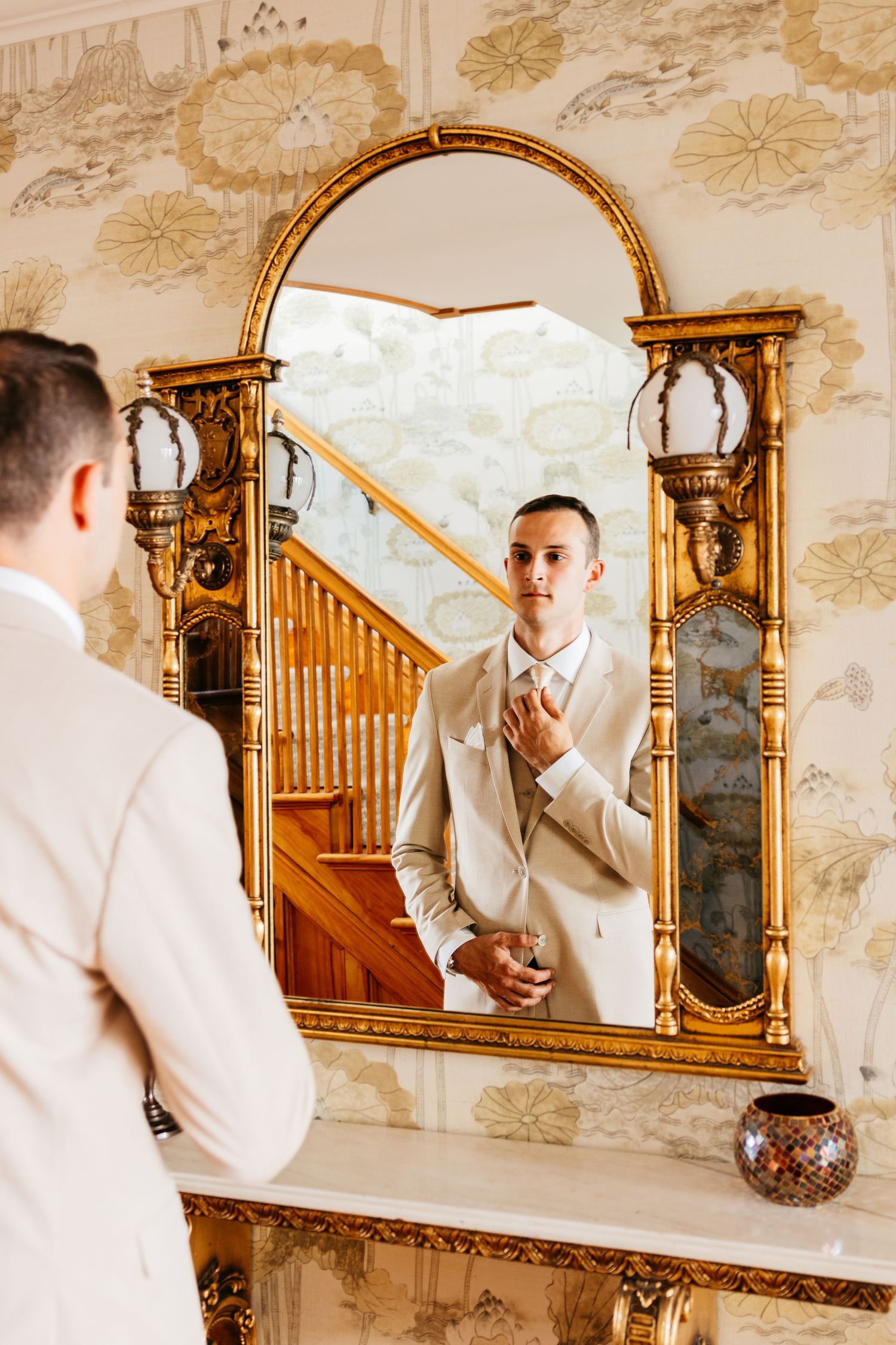 A series of elegant mirror reflections show a groom in a light-colored suit adjusting his tie in an ornate gilded frame.