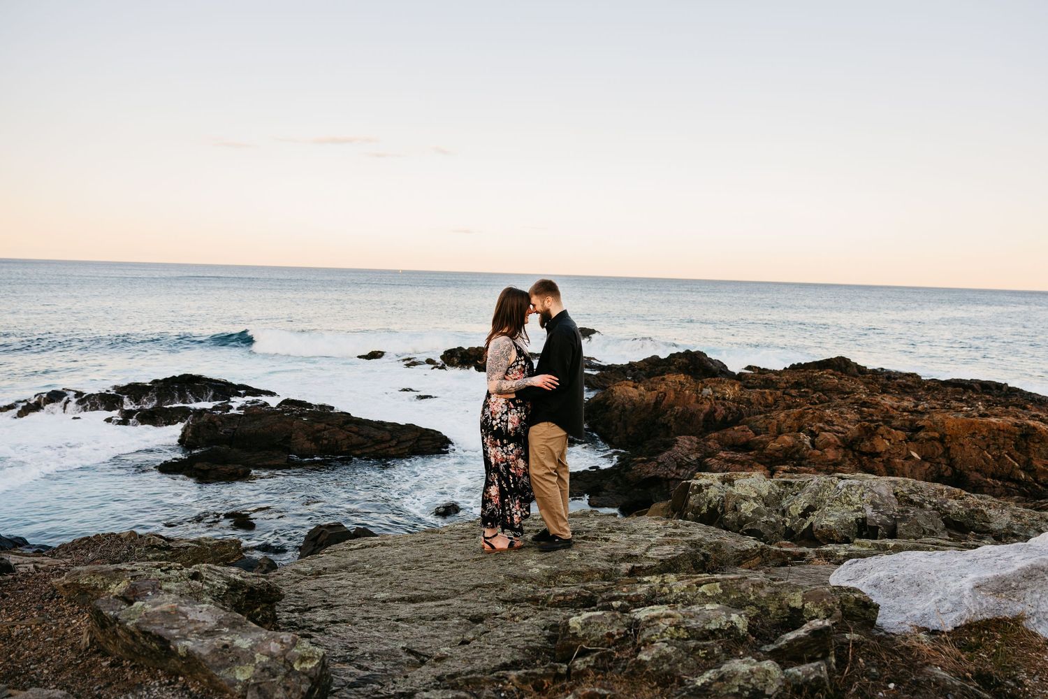Couple embracing on rocky coastal cliffs at sunset with ocean waves in the background.