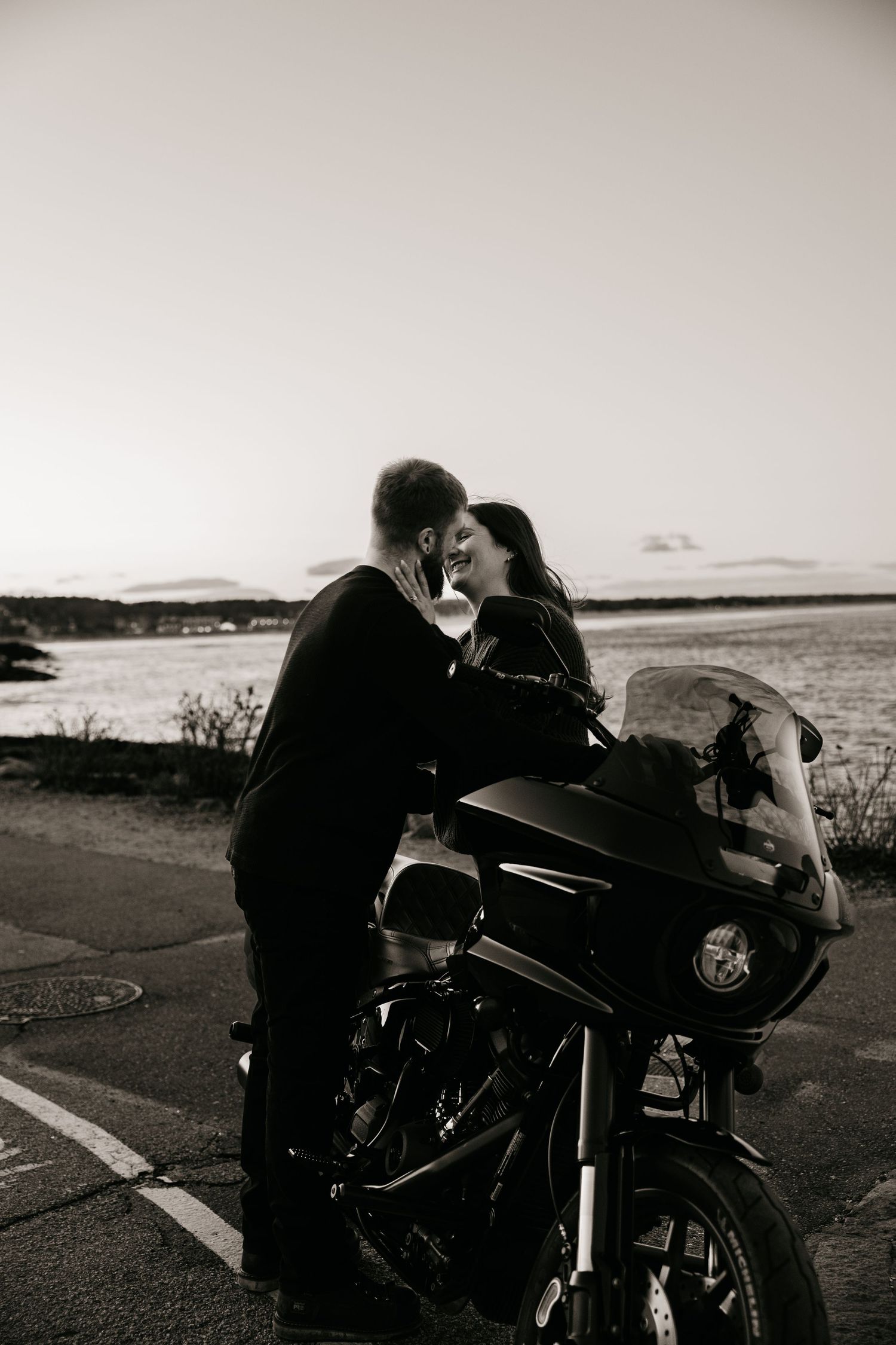 Black and white photo of a romantic couple leaning in for a kiss beside a motorcycle near a scenic waterfront.