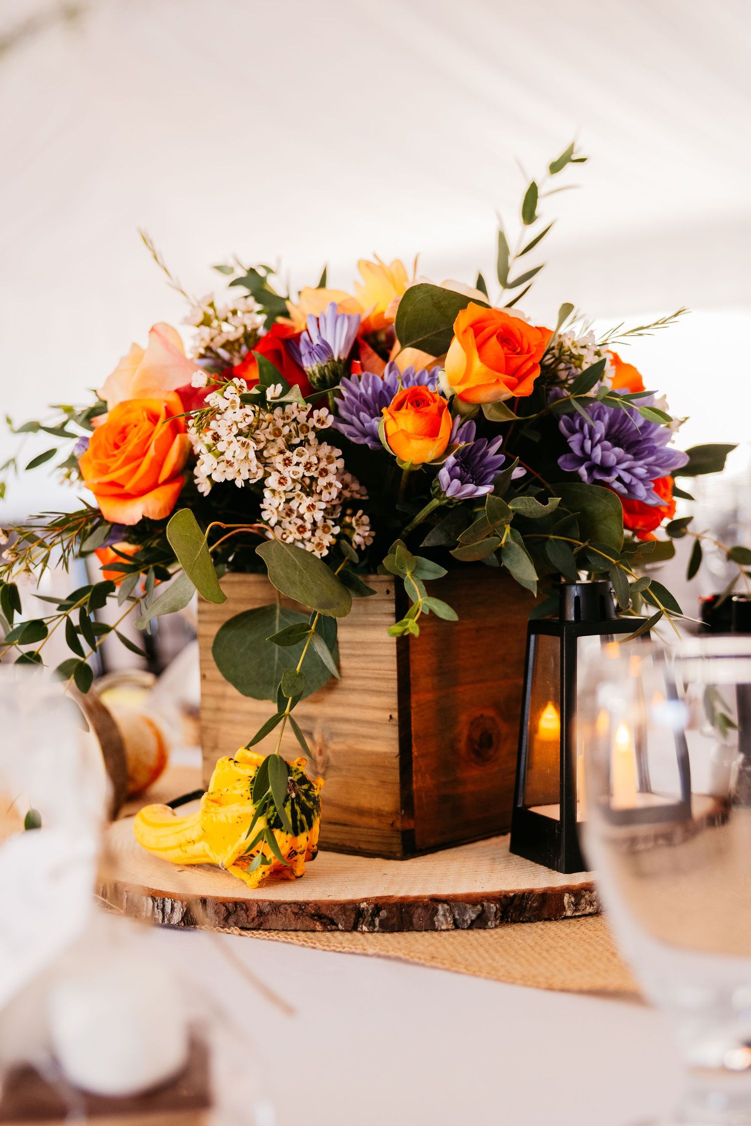 Colorful floral centerpieces with orange roses and purple flowers arranged in rustic wooden boxes on white tablecloths.