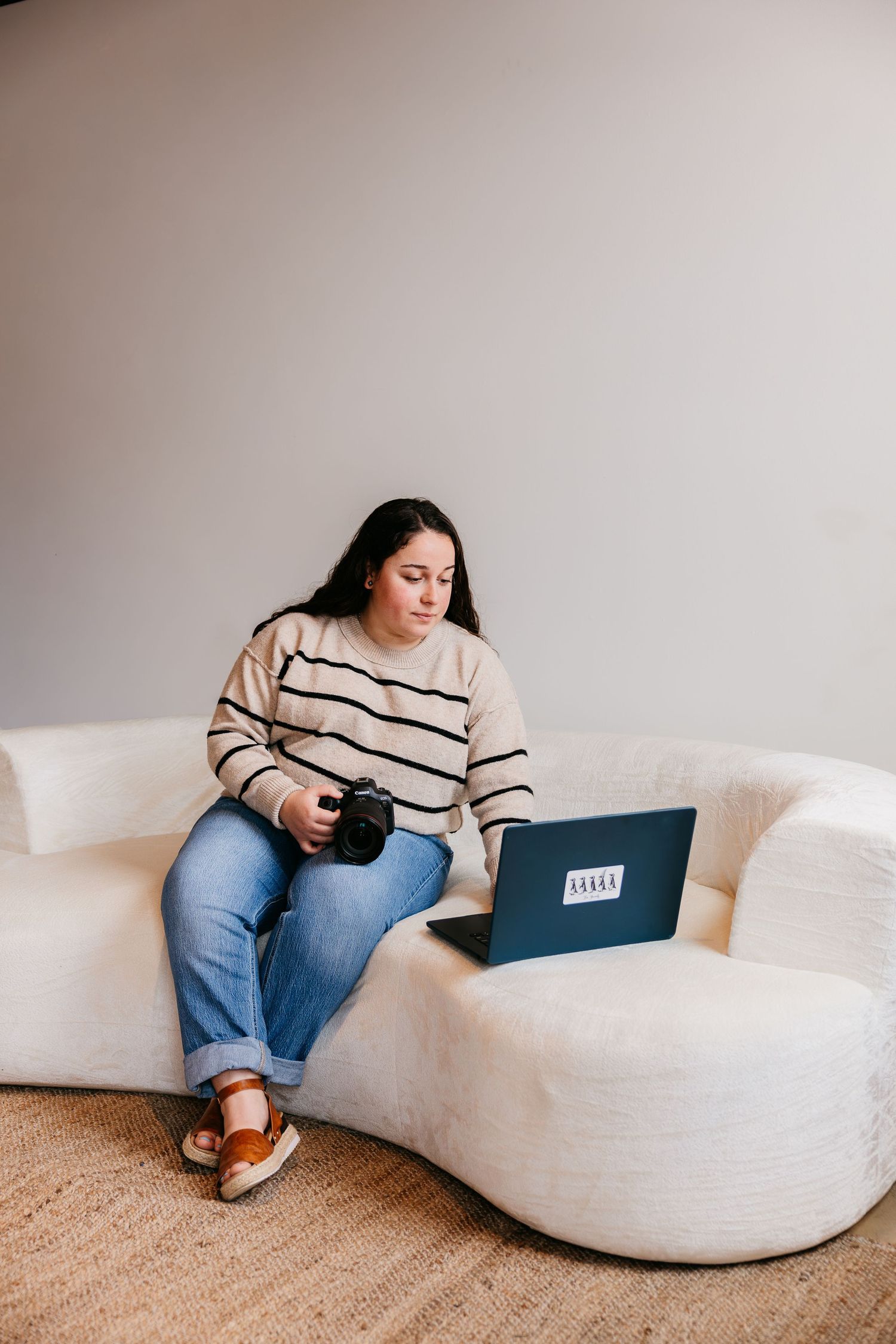 Person in striped sweater and jeans sits casually on a curved white sofa while using a laptop.