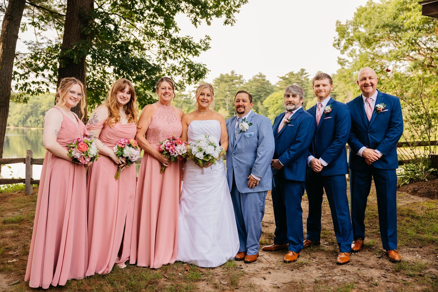 Wedding party posing outdoors, bridesmaids in pink gowns and groomsmen in navy suits flanking bride and groom near a lake.