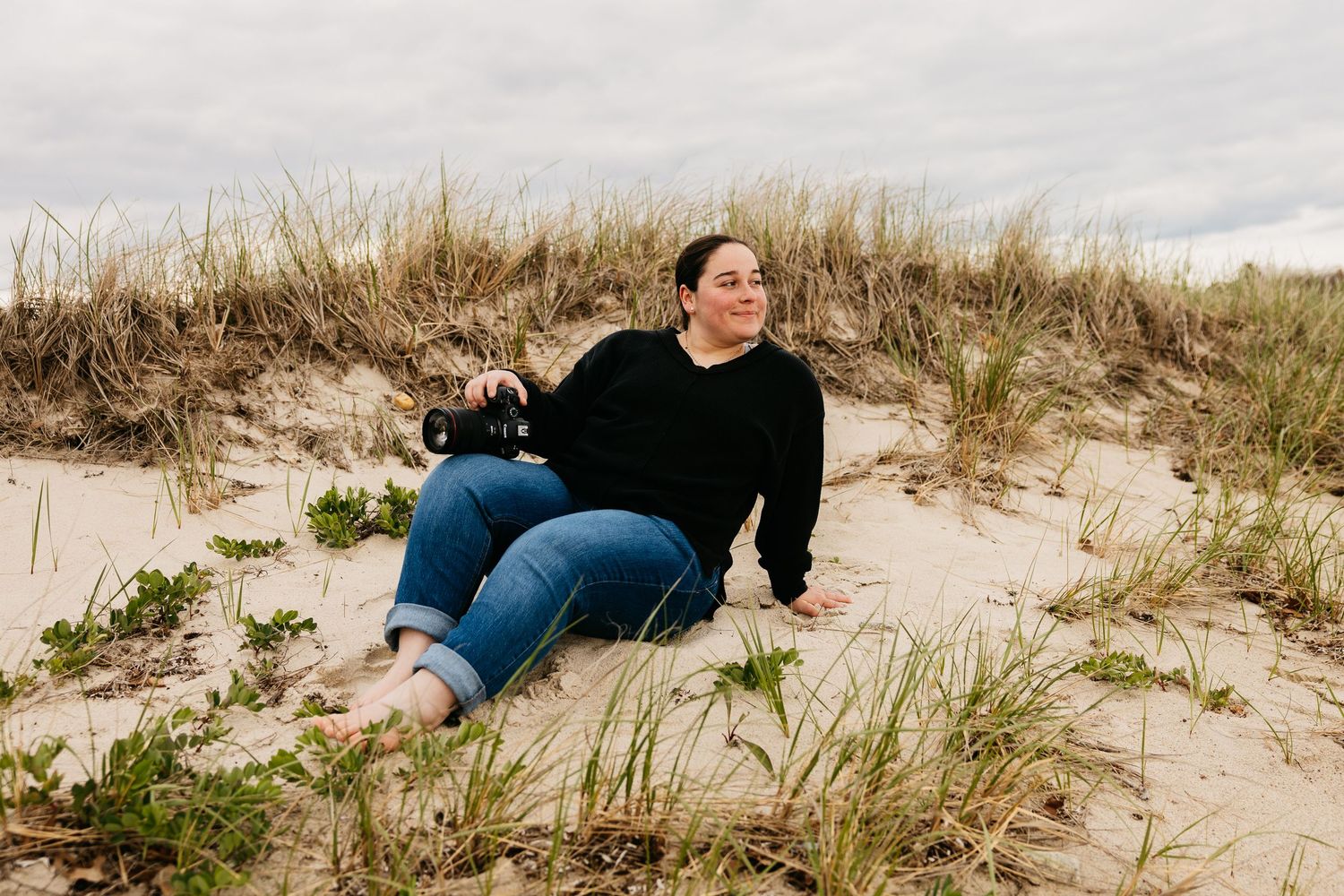 Woman in black sweater and jeans sitting barefoot on sandy beach dunes with tall grass under cloudy sky.