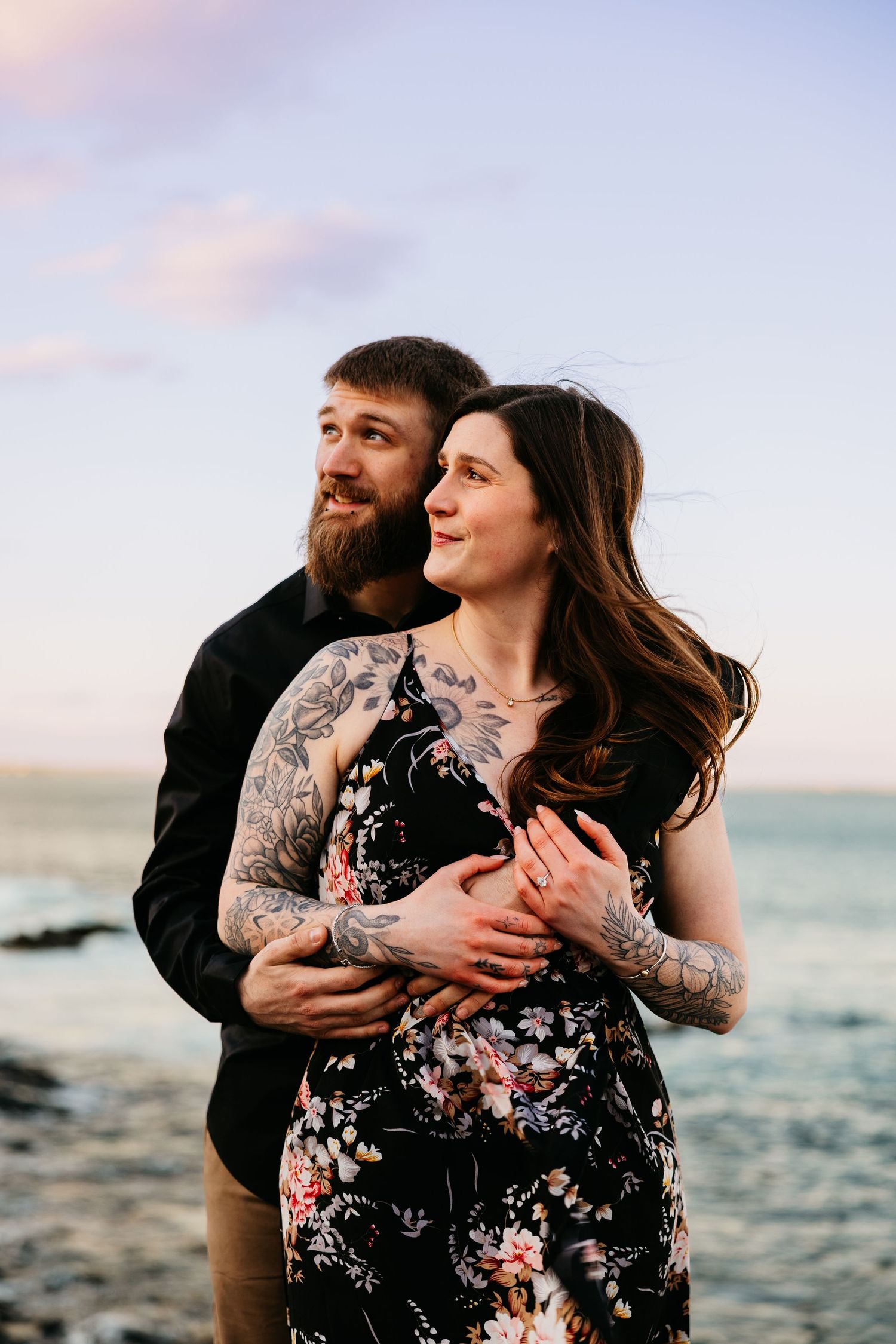 A couple embraces on a rocky beach at sunset, the woman in a floral dress with tattoos, the man in a black shirt with a beard.