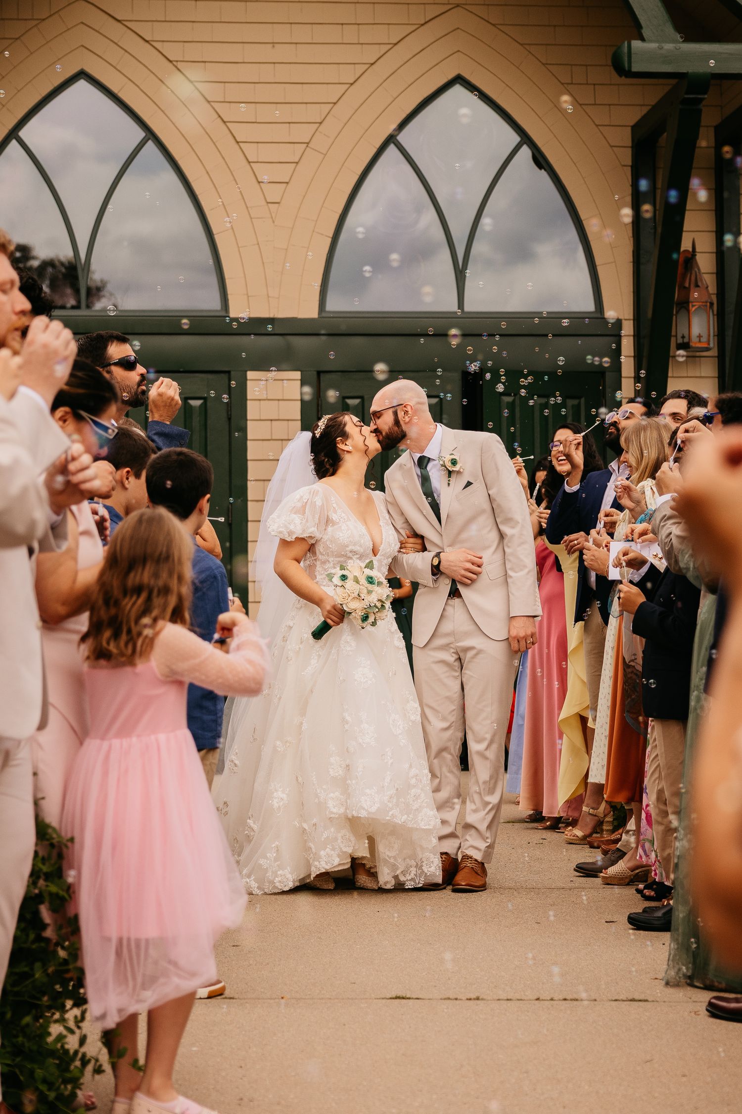 Bride and groom share a kiss during their wedding exit surrounded by bubbles and cheering guests outside a chapel.