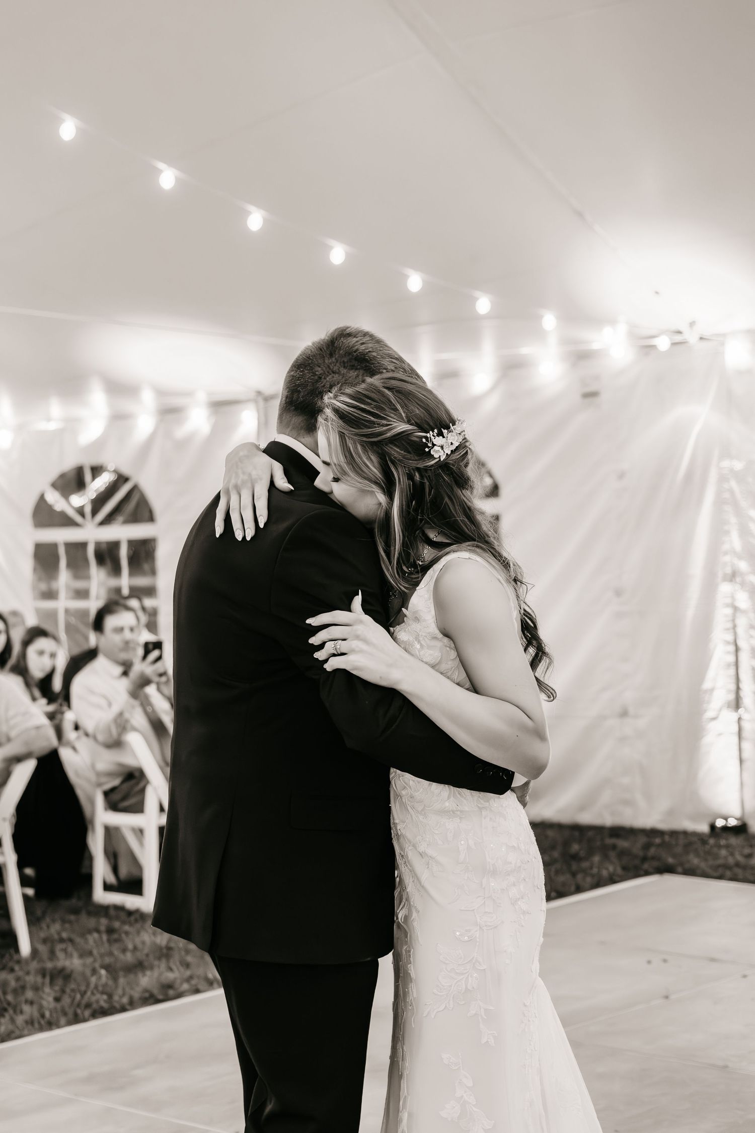 A romantic first dance sequence captured in black and white under a wedding tent with string lights overhead.