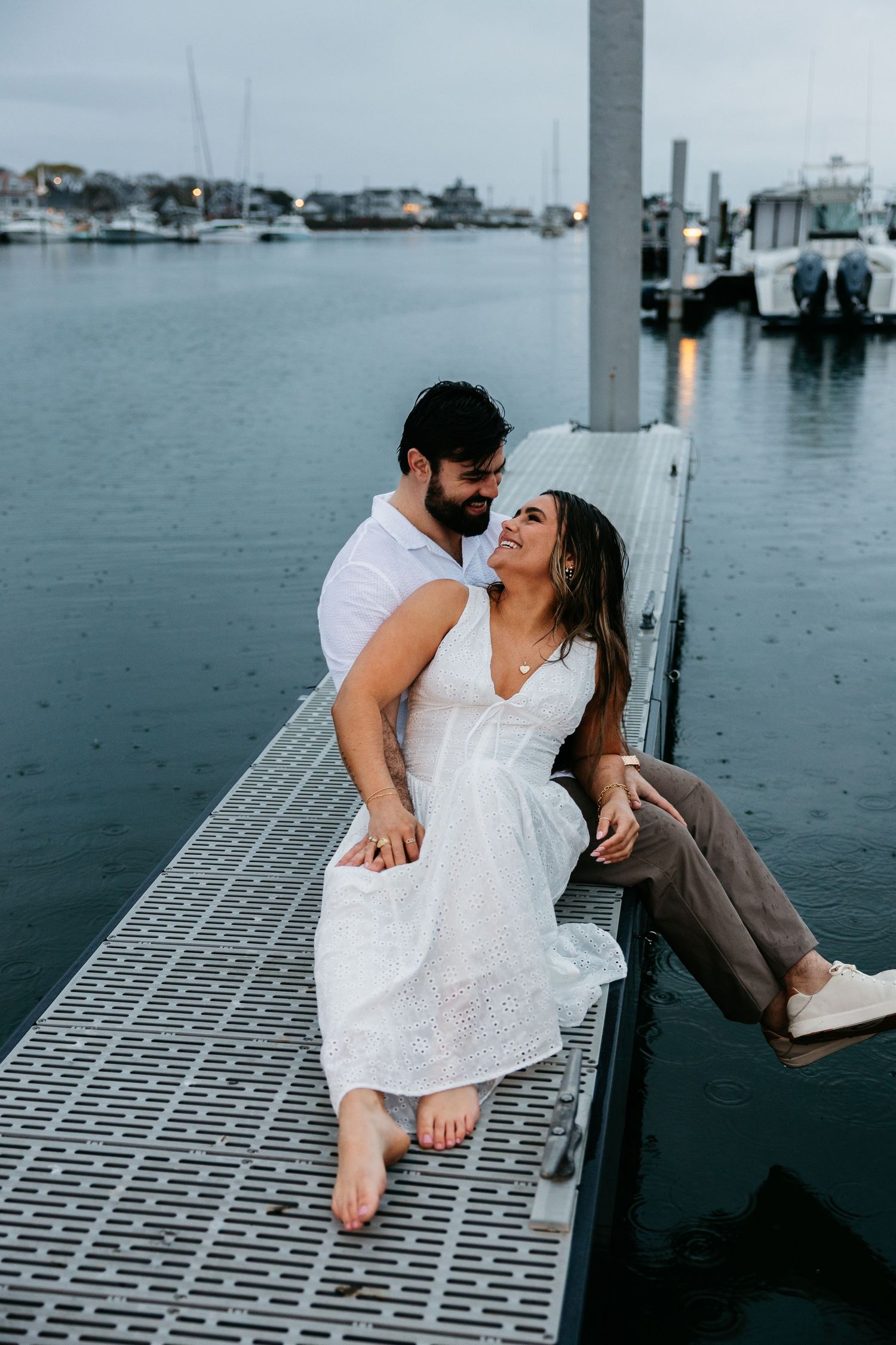 Couple sitting on a marina dock at dusk, woman in white dress, man in white shirt, with boats and water in background.