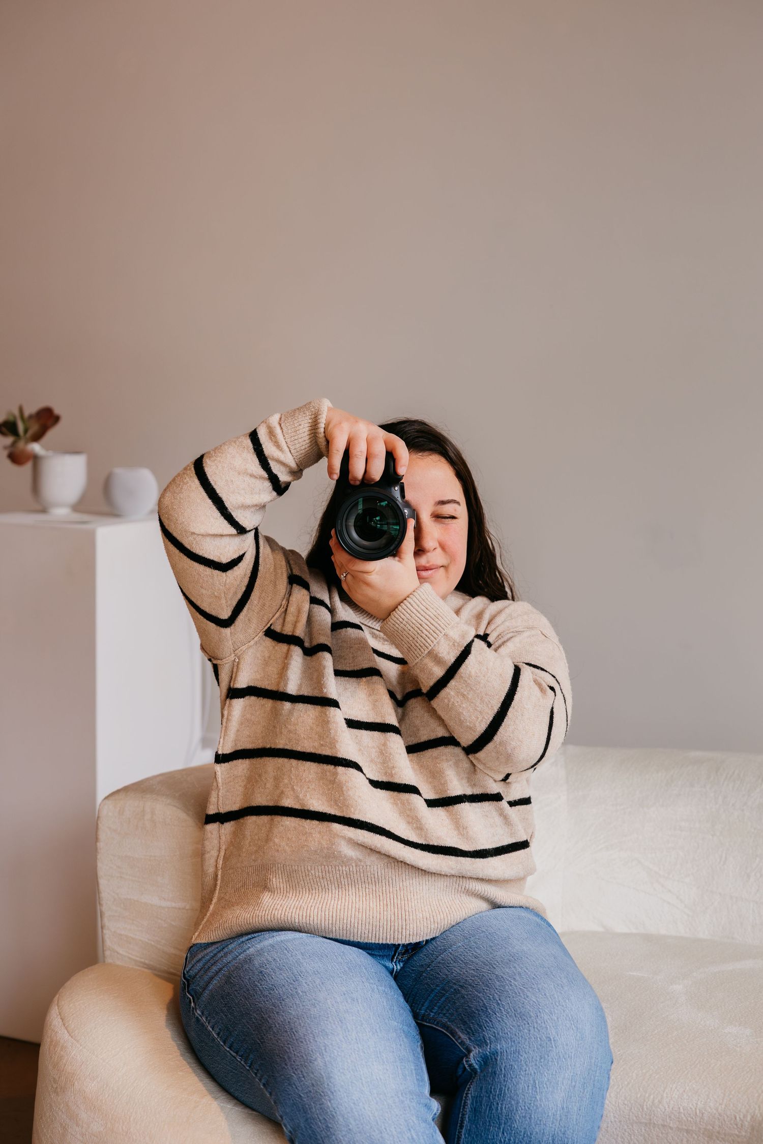 Person wearing beige striped sweater and blue jeans sits on white couch taking photos with camera.